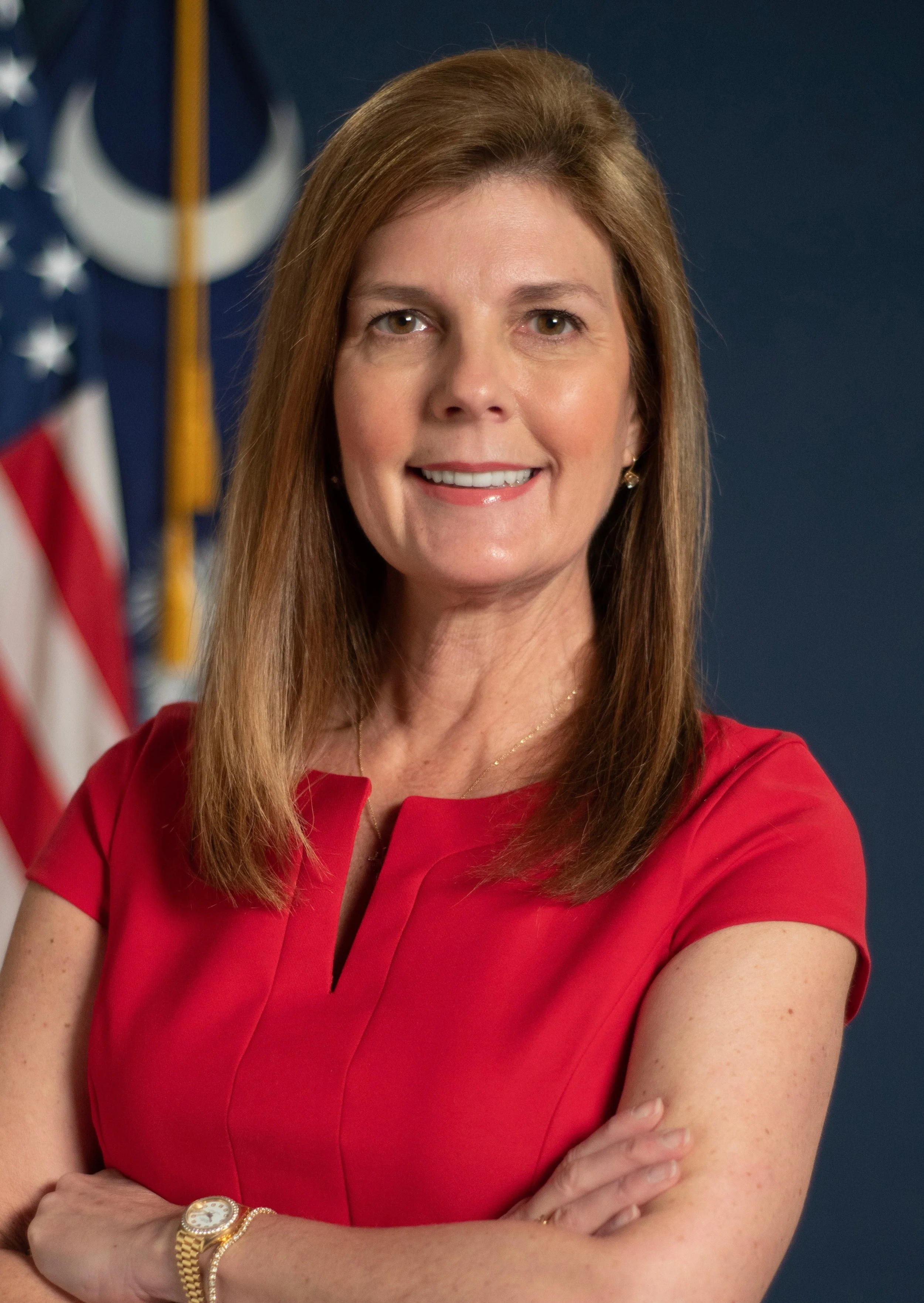 A woman with shoulder-length brown hair wearing a red dress, arms crossed, smiling, with American and military flags in the background.