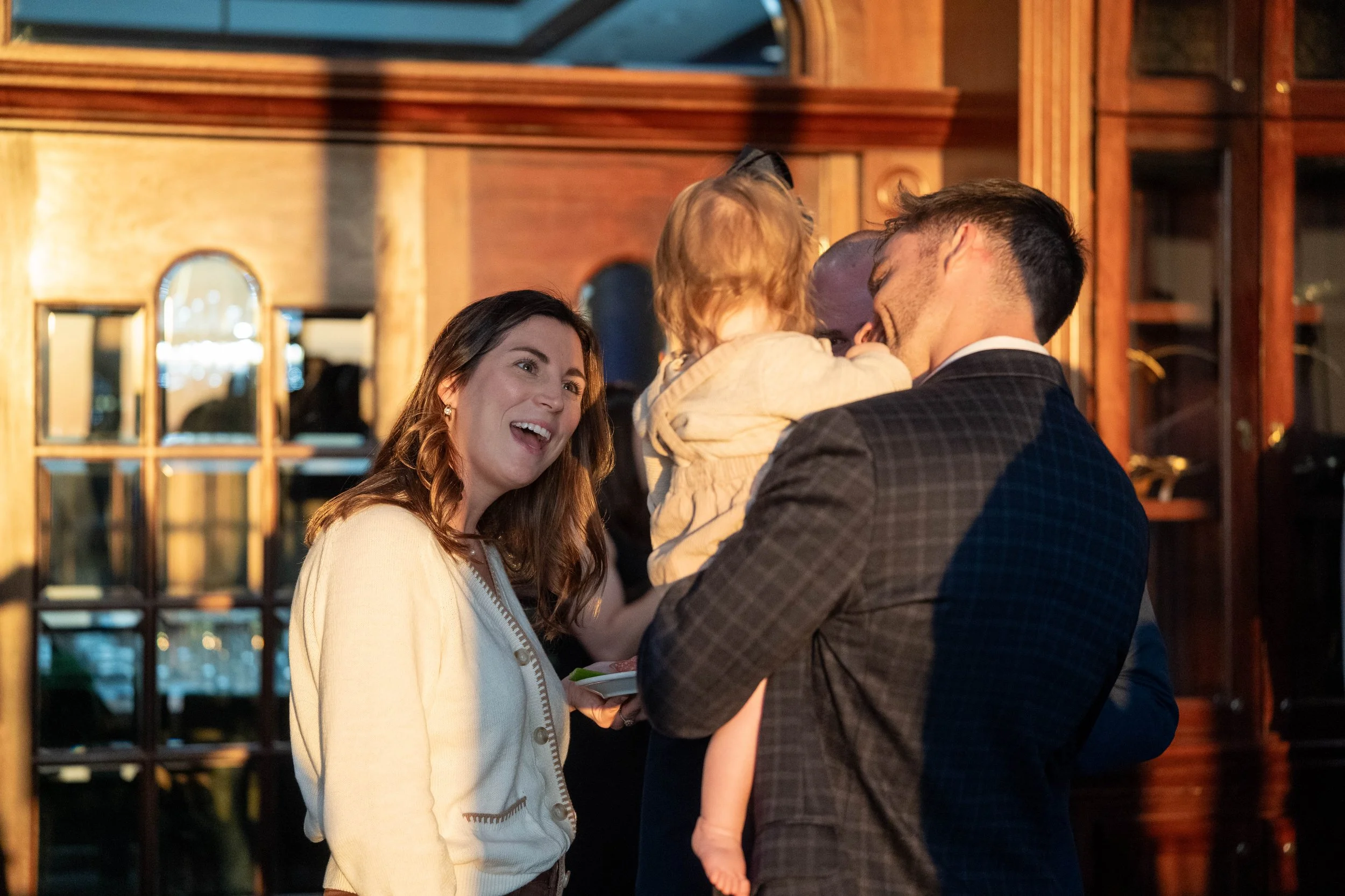 A woman is smiling and talking to a man holding a child at a social gathering indoors, with wooden furniture and warm lighting.
