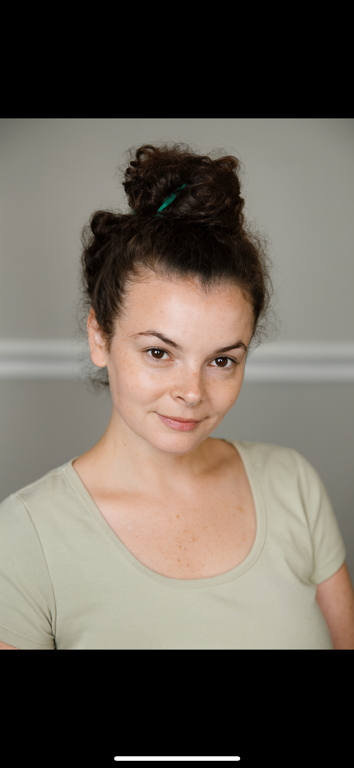A young woman with her dark brown, curly hair tied up in a messy bun, wearing a light beige t-shirt, smiling gently, with freckles on her face, posing against a gray background.