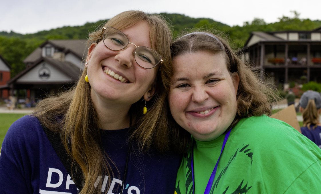 Two smiling women hugging outdoors, with houses and a green hill in the background.