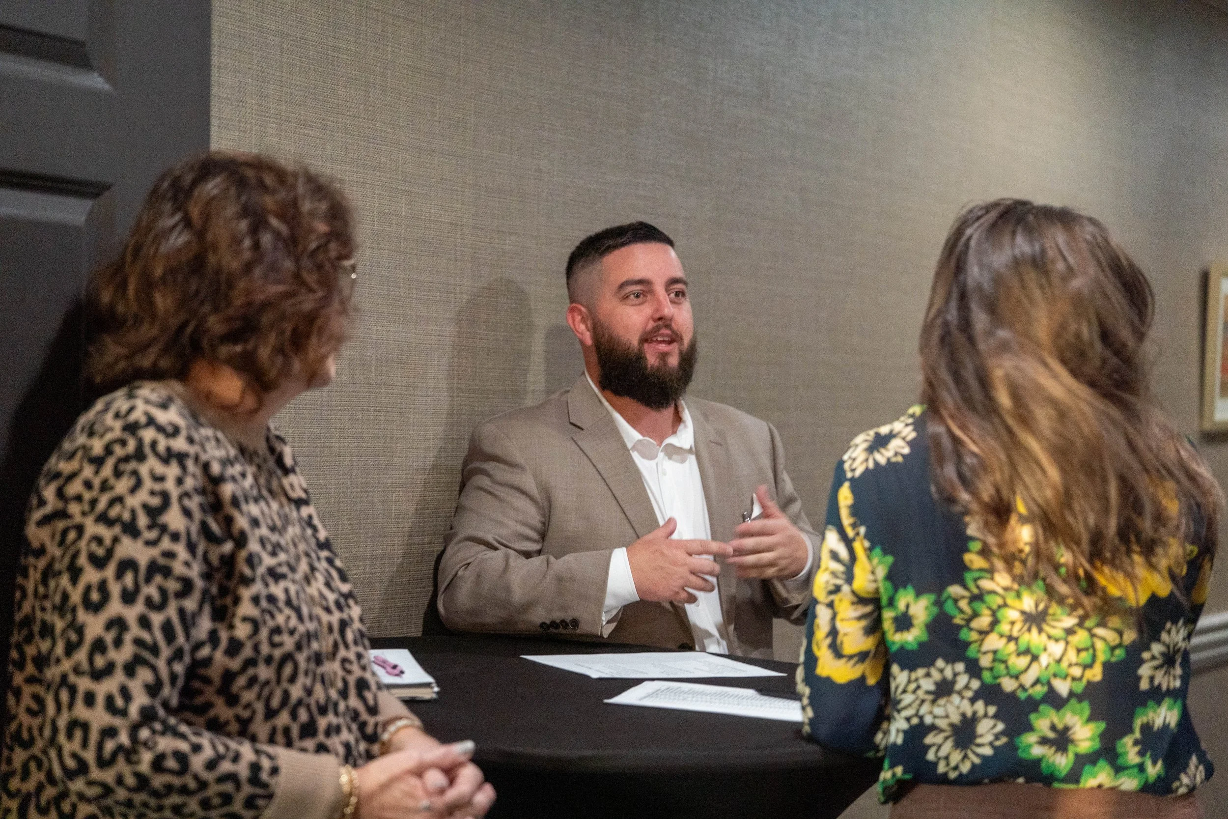 A man in a beige suit speaking to two women at a table during a discussion or interview, with a third woman partially visible on the left.