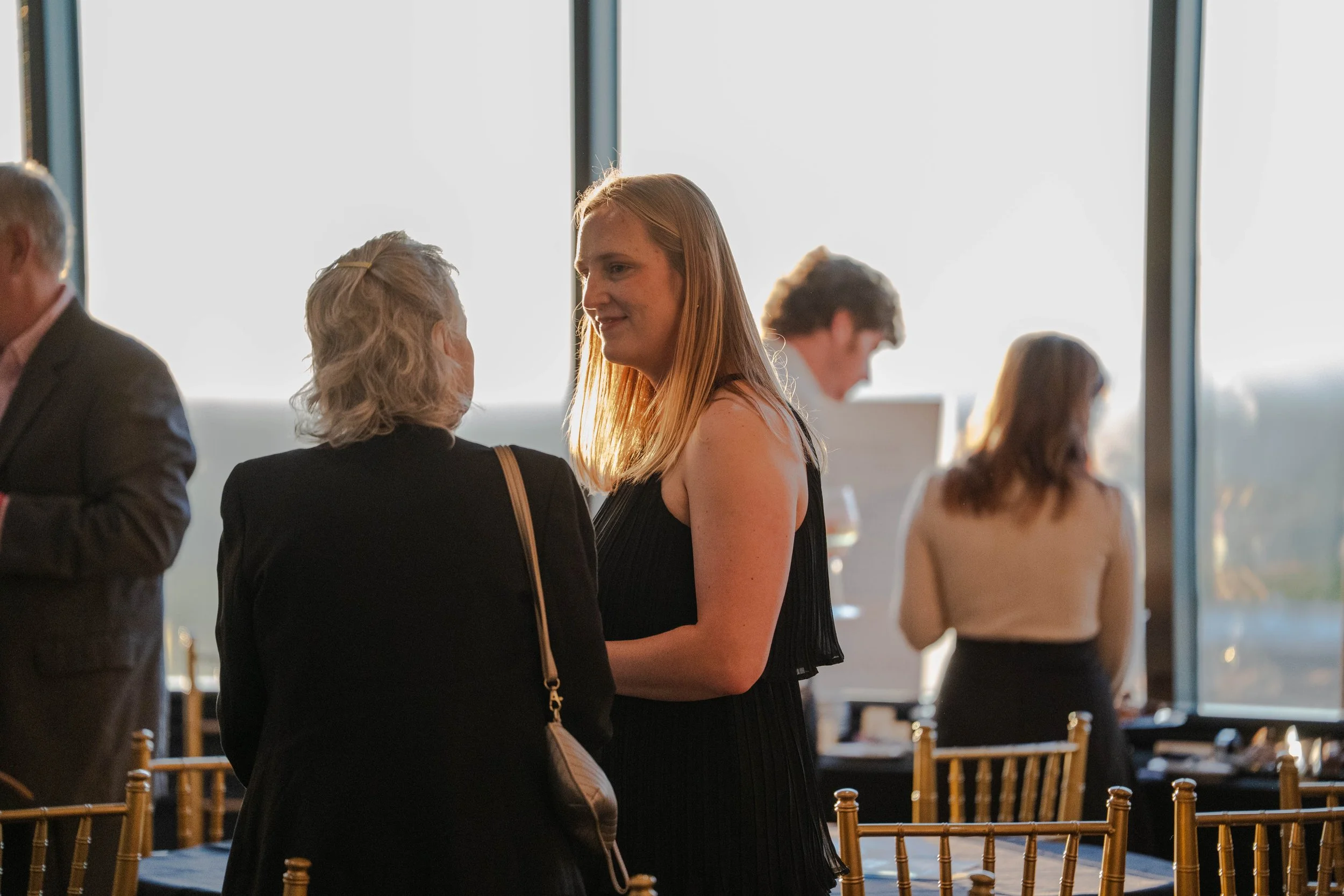 People at a social gathering inside a room with large windows, engaging in conversations, with some standing and others sitting near tables and chairs.