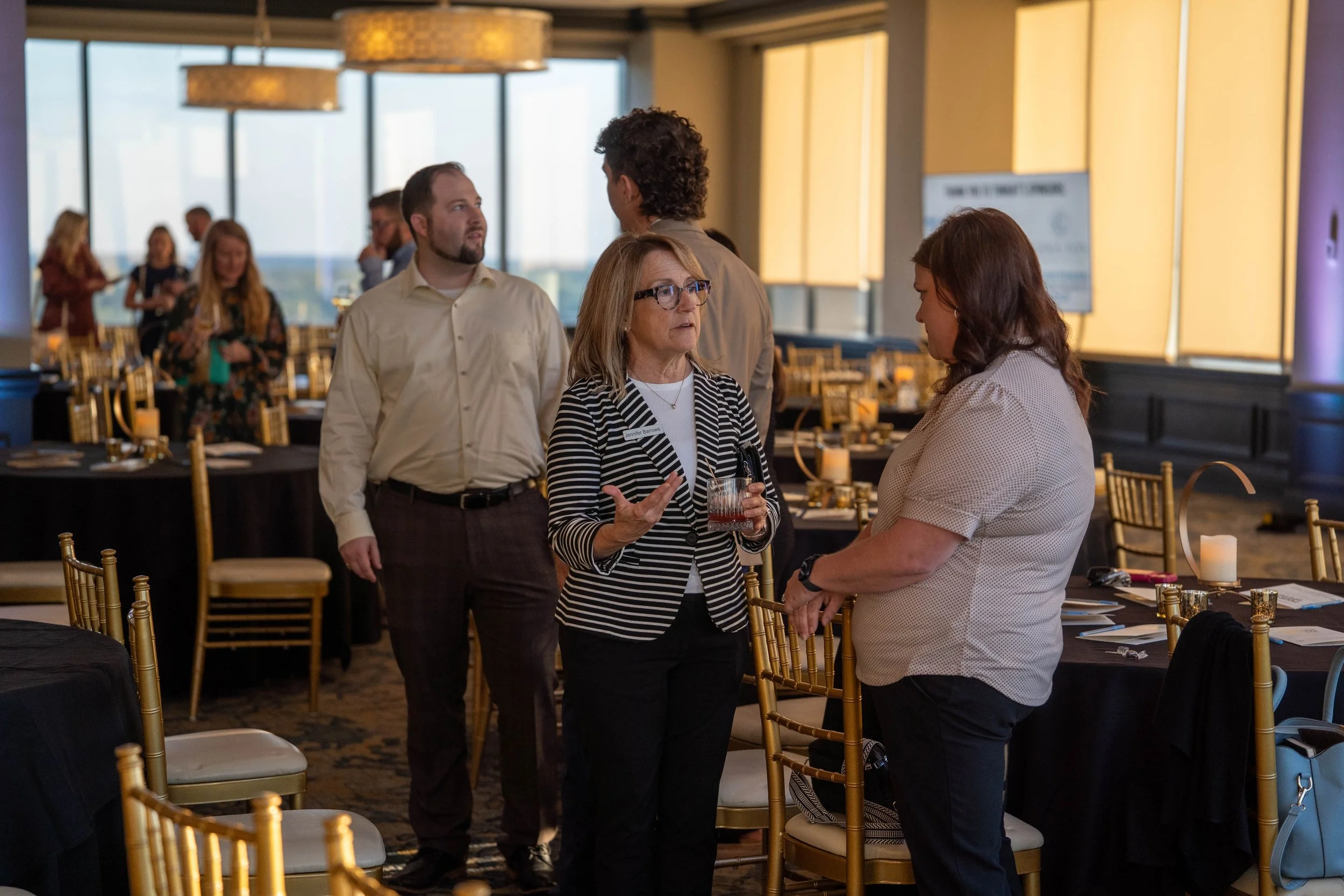 Three women and two men conversing in a banquet hall with gold chairs and high windows in the background.
