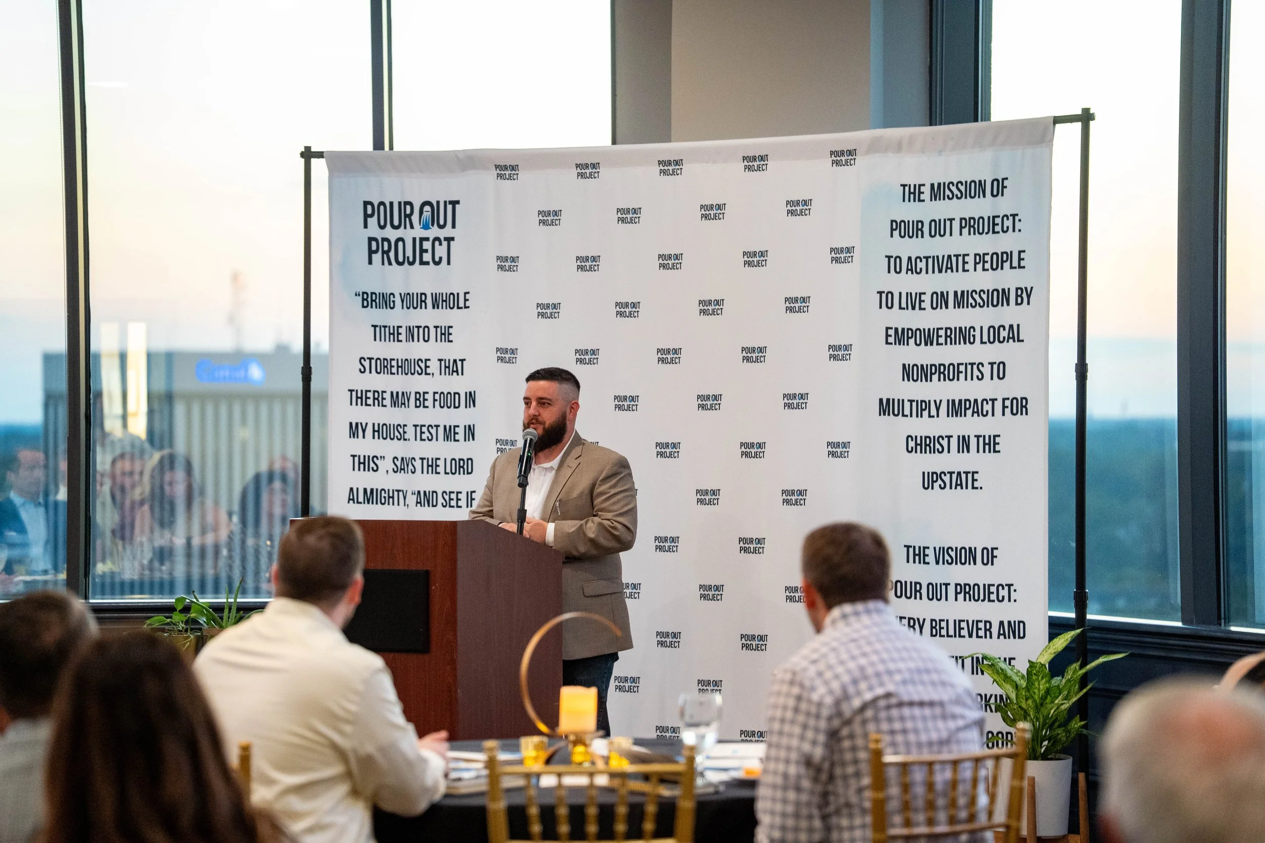 Man in beige suit speaking at a podium during a conference, with a banner behind him that has text about the Pour Out Project and a quote from the Bible, in a room with large windows and an audience.