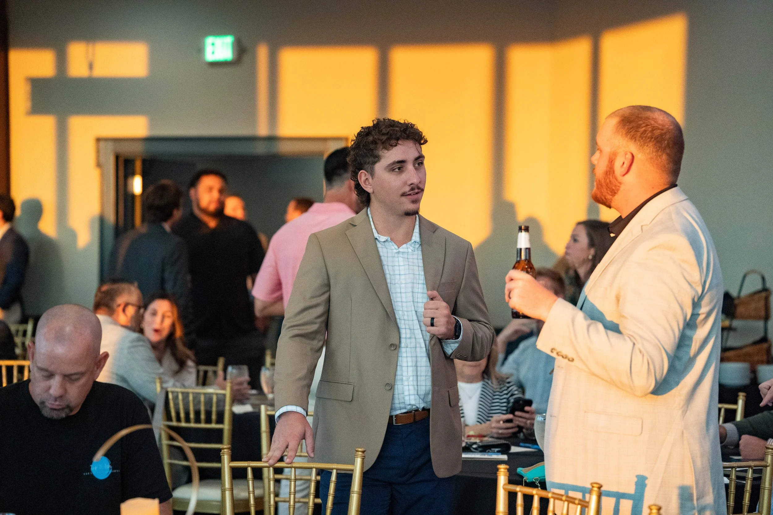 Two men in suits having a conversation at a social event, one holding a beer, with other people seated and mingling in the background.