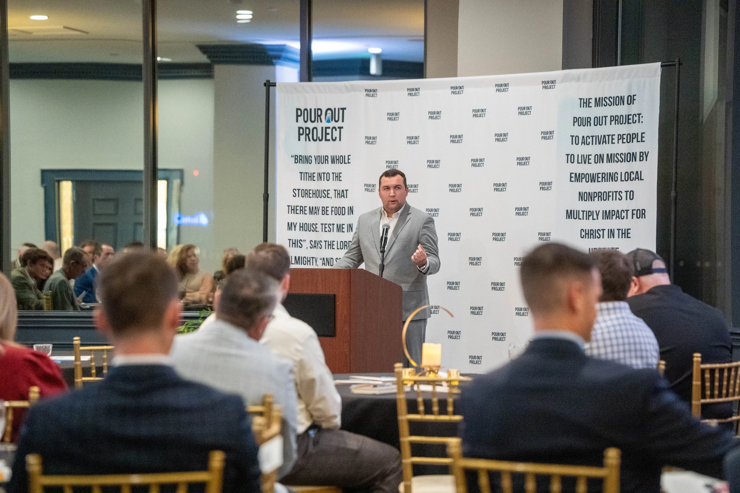 A man in a gray suit speaking at a podium during a conference with a large audience. There is a patterned white banner behind him with the title "Pour Out Project" and text explaining the mission to activate people to live on mission by empowering local nonprofits.