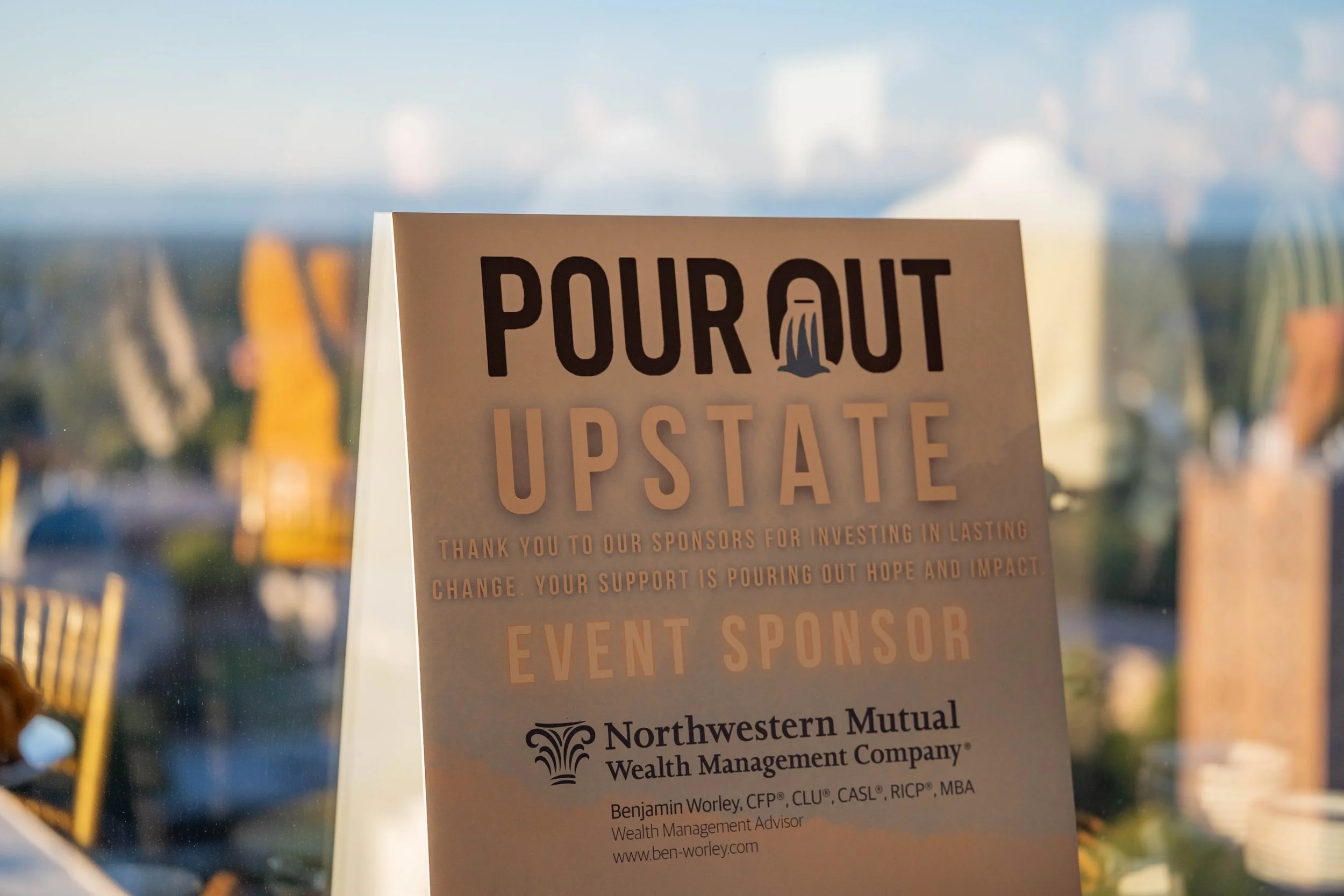 Event sign with the text 'Pour Out Upstate' thanking sponsors, featuring Northwestern Mutual Wealth Management Company as the event sponsor, with a cityscape showing through a window in the background.