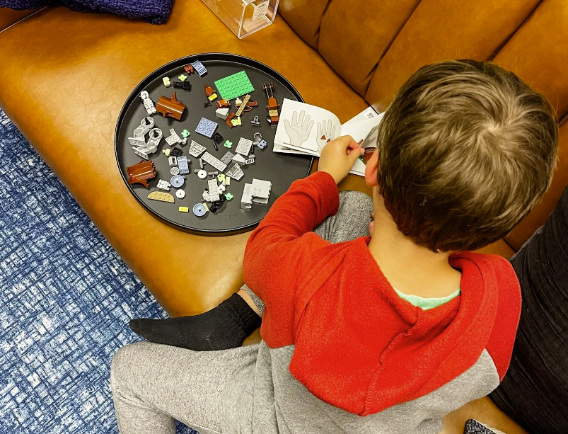 A boy assembling LEGO pieces on a black tray, sitting on a brown leather couch with a book open in front of him.