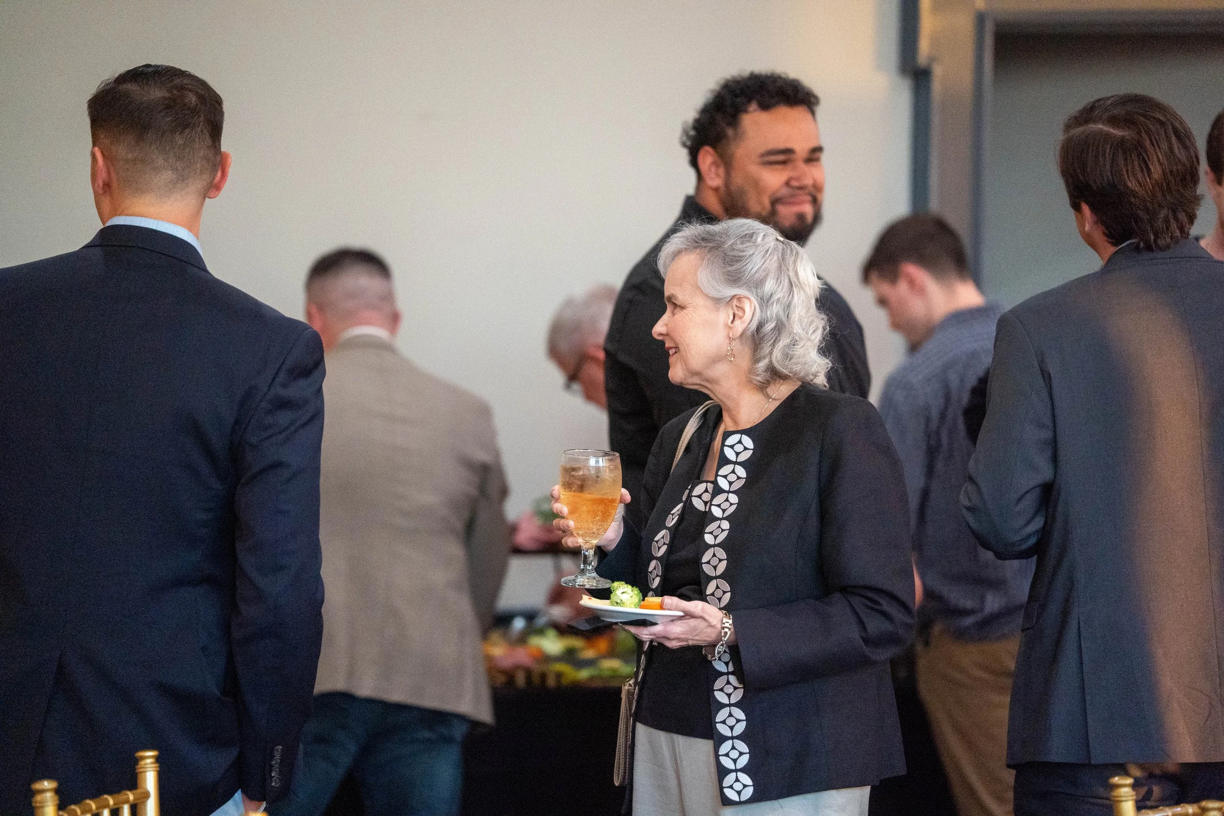 A woman with gray hair holding a glass of wine and a plate of snacks at a social gathering, smiling and talking to a man in a dark suit, surrounded by other people in business attire.