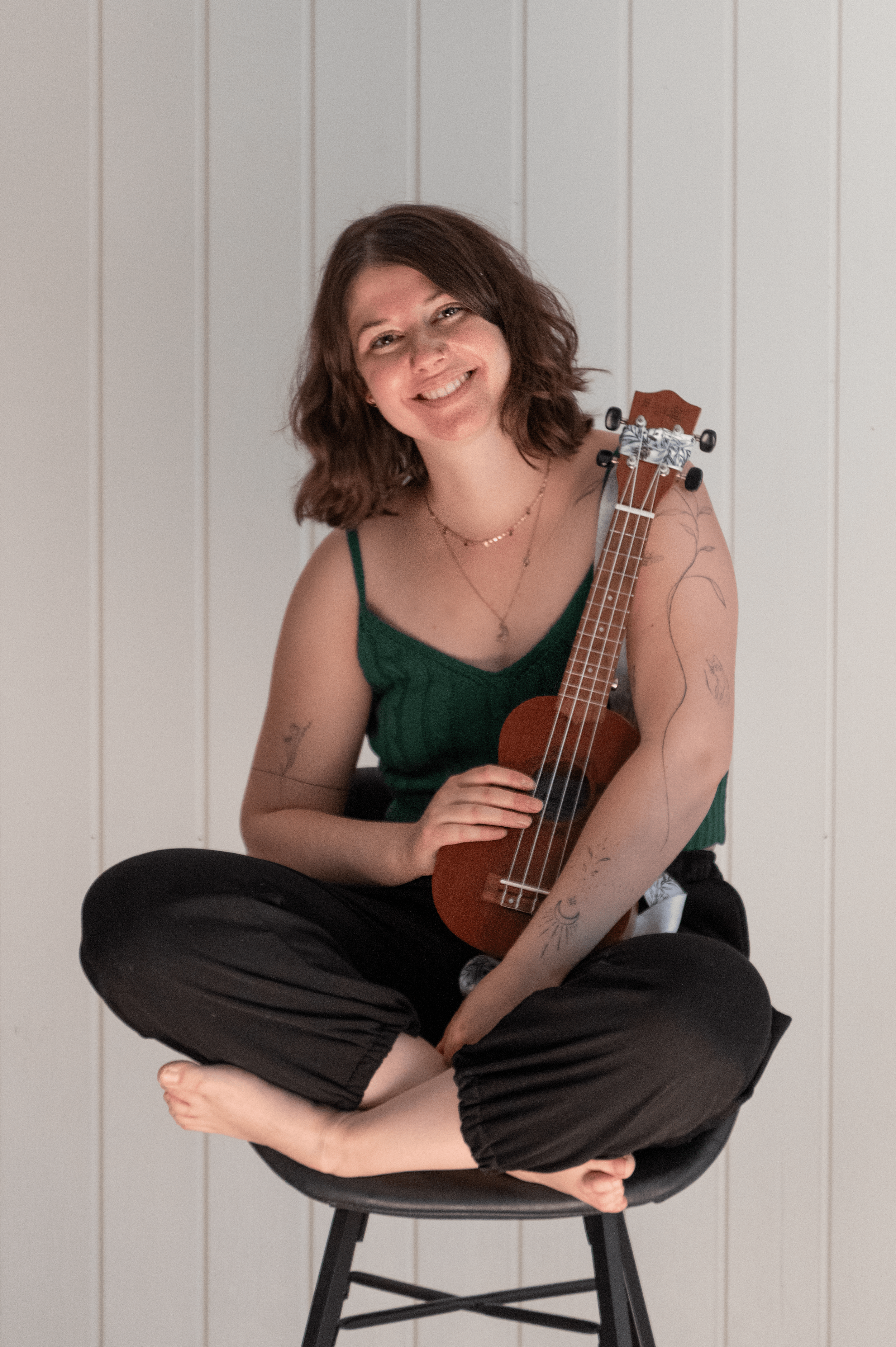 Eine junge Frau mit braunen, lockigen Haaren sitzt barfuß auf einem Stuhl und hält eine Ukulele. Sie trägt ein grünes Top und schwarze Hosen, hat Tattoos an den Armen und trägt mehrere Halsketten, während sie lächelnd in die Kamera schaut vor einer weißen Wand.