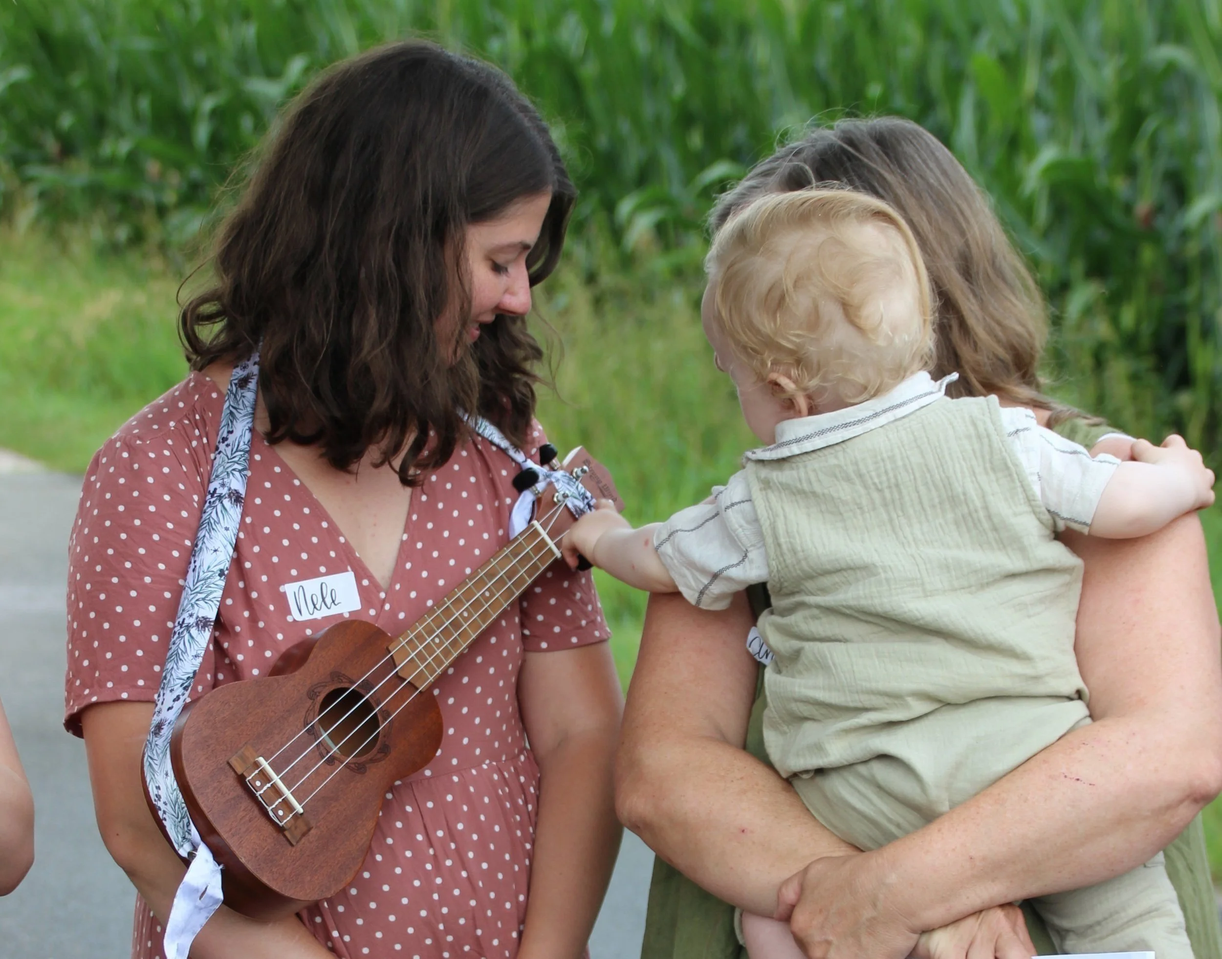 Drei Menschen, eine junge Frau mit einem Ukulele, ein Baby und eine weitere erwachsene Frau, auf einer Wiese mit hohen grünen Pflanzen im Hintergrund, interagieren freundlich miteinander.