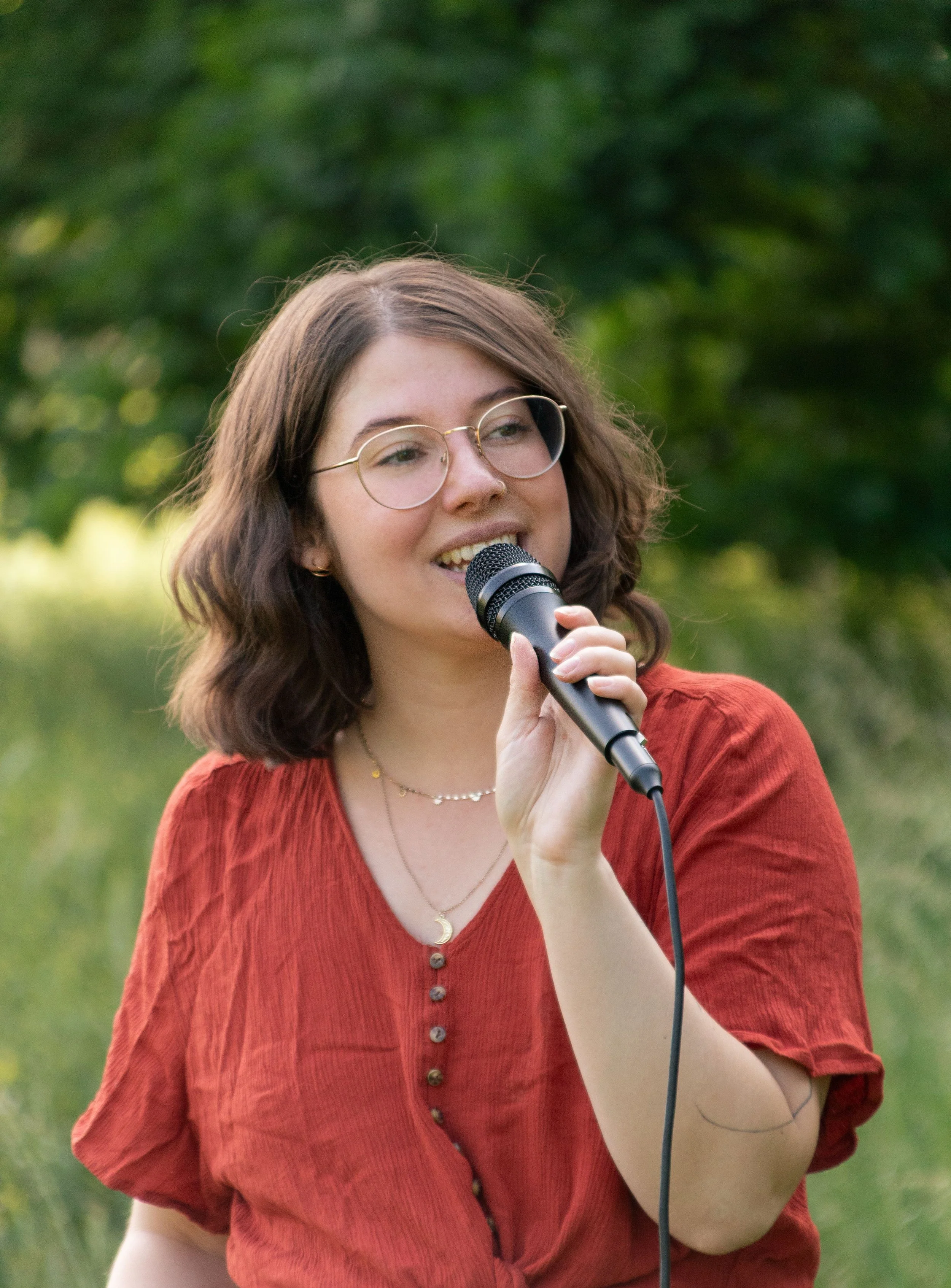 Eine junge Frau mit Brille und braunen, lockigen Haaren hält eine Handmikrofon im Freien, umgeben von Bäumen und grüner Natur.