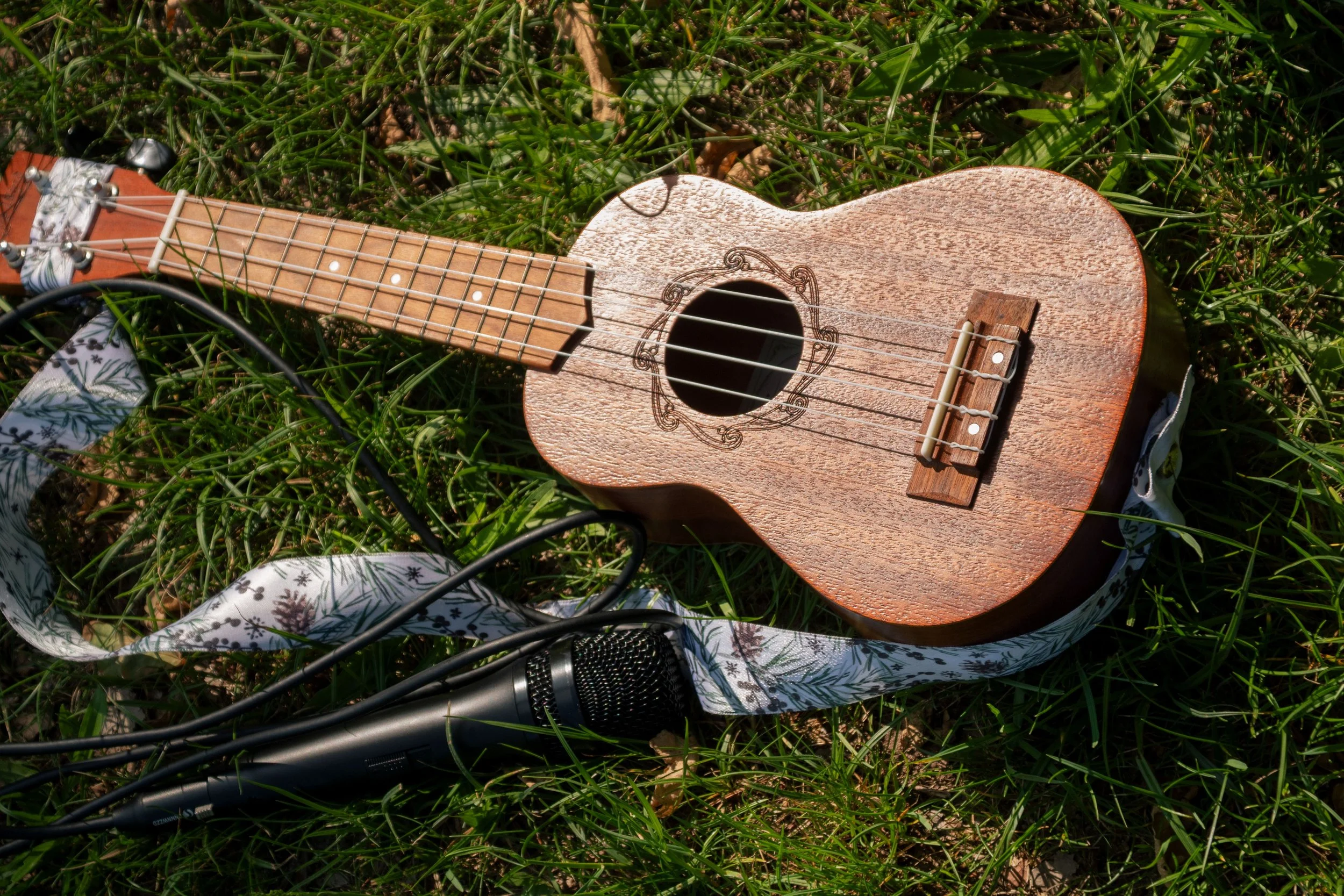 Eine kleine Holzgitarre mit verzierten Korpus liegt auf grünem Gras, verbunden mit einem Mikrofon und einem Kabel.