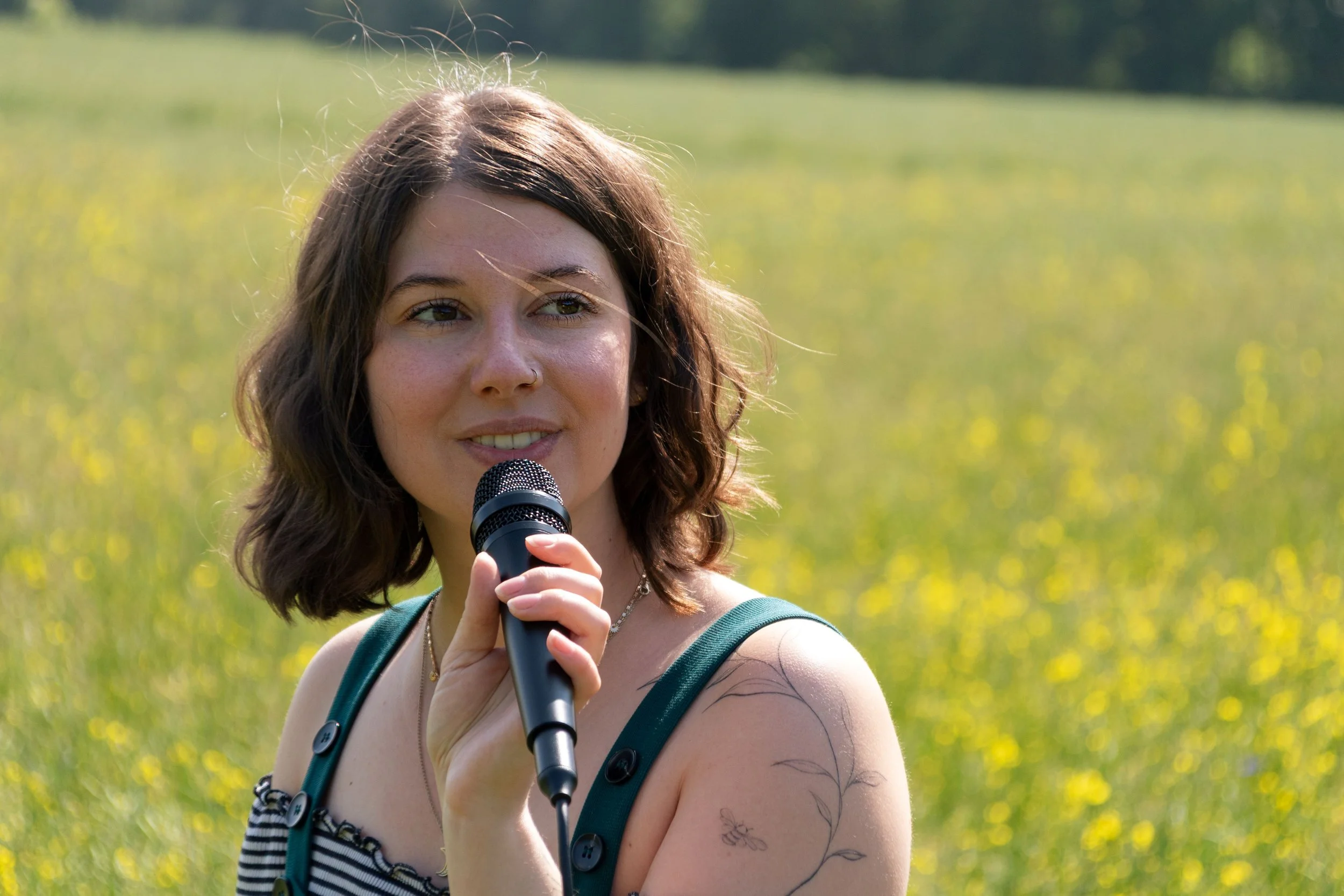 Junge Frau mit kurzen braunen Haaren, Sonnenbrille auf dem Kopf, hält ein Mikrofon und steht in einem grünen Feld, sonniges Wetter.