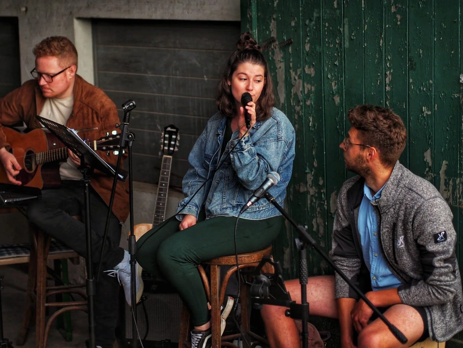 Drei junge Menschen, zwei Männer und eine Frau, in einem Musik-Setup. Der Mann auf der linken Seite spielt Gitarre, die Frau in der Mitte singt mit einem Mikrofon, und der Mann auf der rechten Seite hört aufmerksam zu. Sie befinden sich in einem Raum mit dunklen Holzwänden.