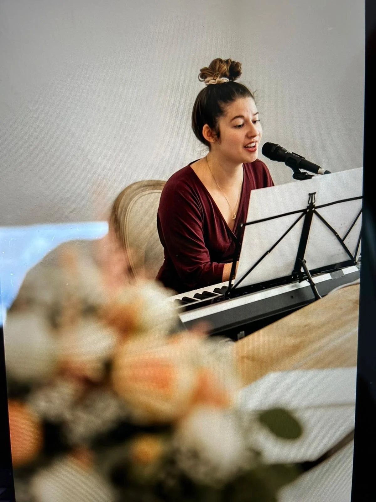 Eine junge Frau singt beim Klavierspielen vor einem Mikrofon, während im Vordergrund Blumen liegen.