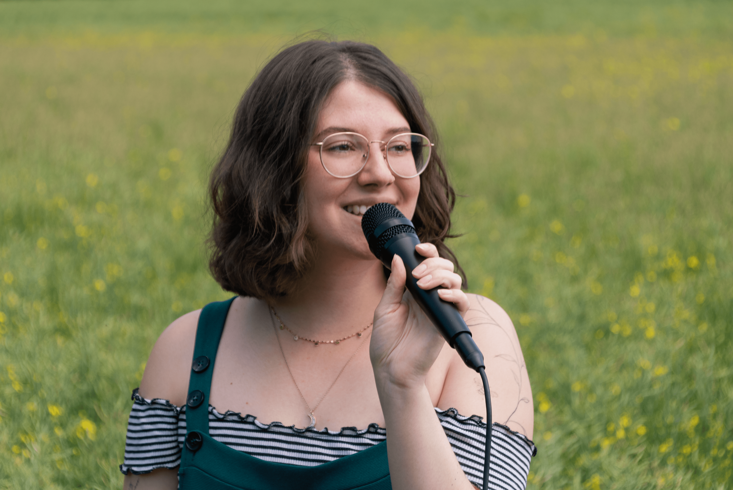 Junge Frau im Grünen mit Mikrofon, trägt Brille und ein ärmelloses Top mit Streifen, im Hintergrund ein Feld mit kleinen gelben Blumen.