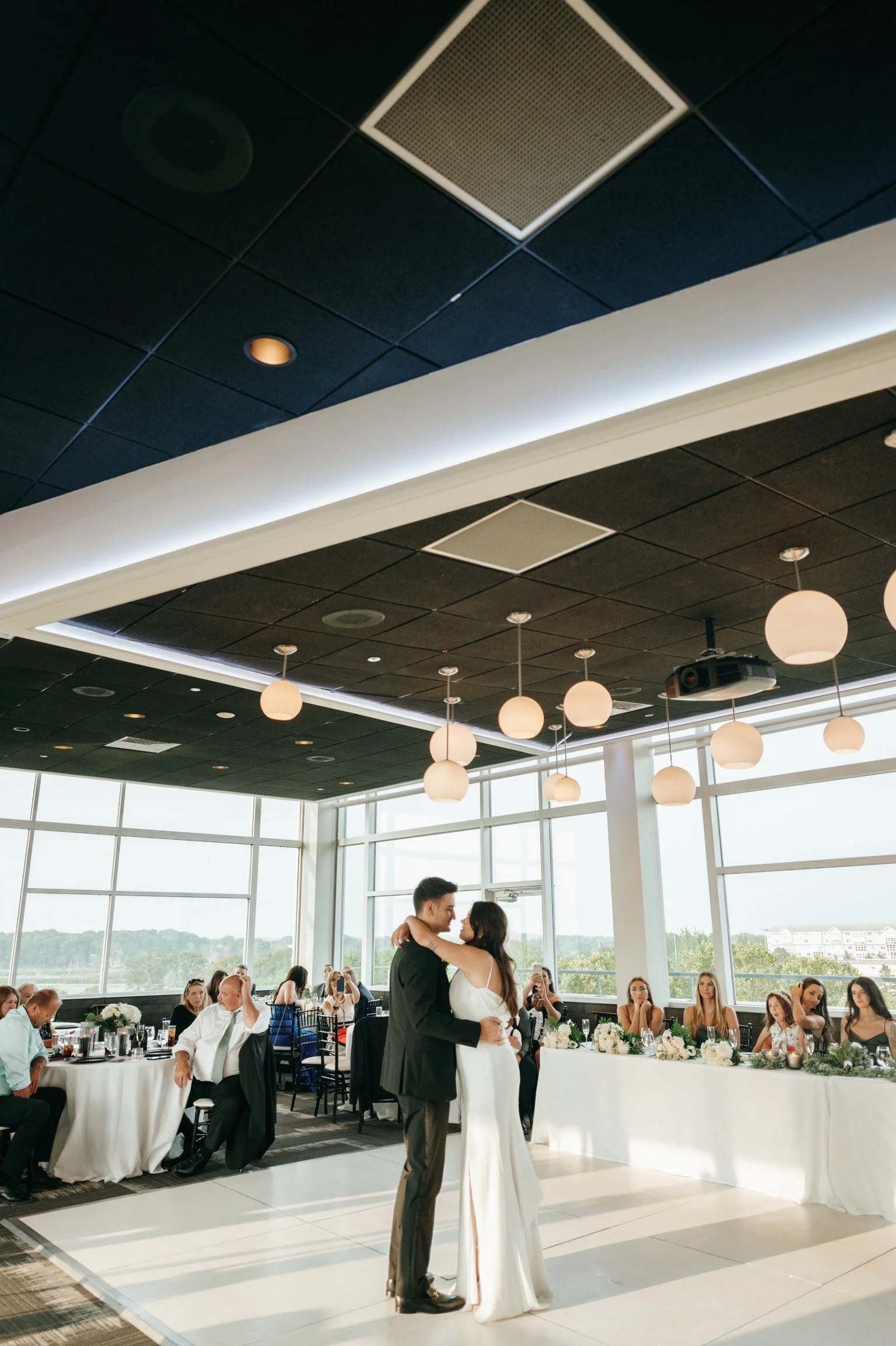 A bride and groom dancing during their wedding reception in a modern venue with large windows and hanging lights, guests seated at tables watching and taking photos.