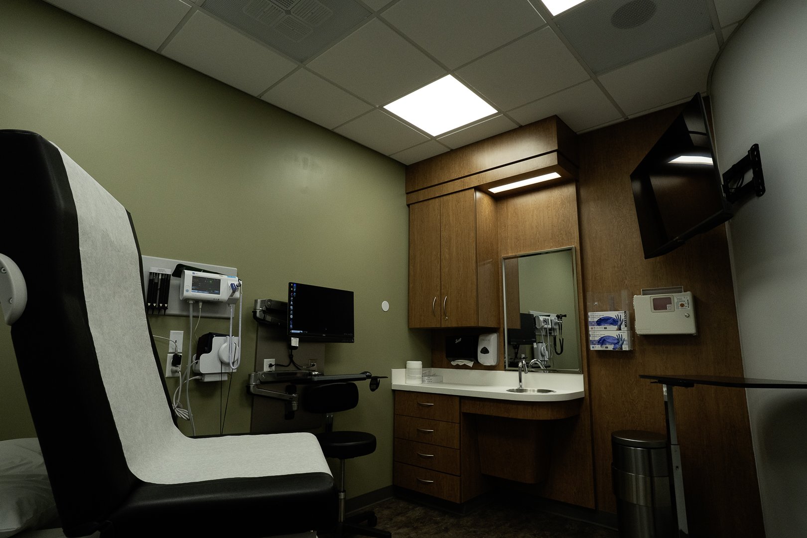 Medical examination room with a hospital bed, medical equipment, a wall-mounted monitor, a sink with a mirror, wood cabinets, a wall-mounted TV, and a trash can.