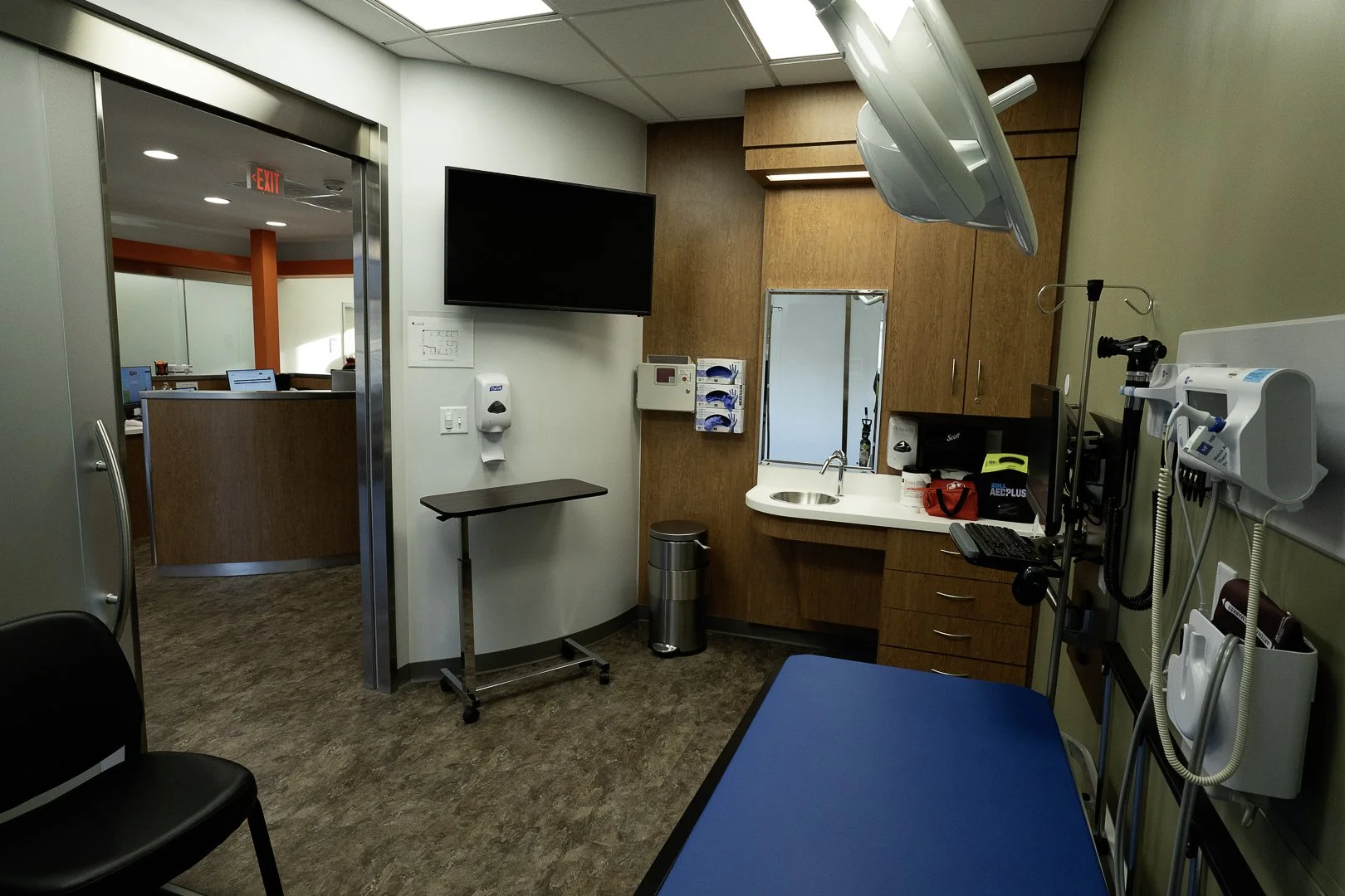 Empty examination room with medical equipment, a blue examination table, a wall-mounted monitor, and a small sink with wood cabinets.