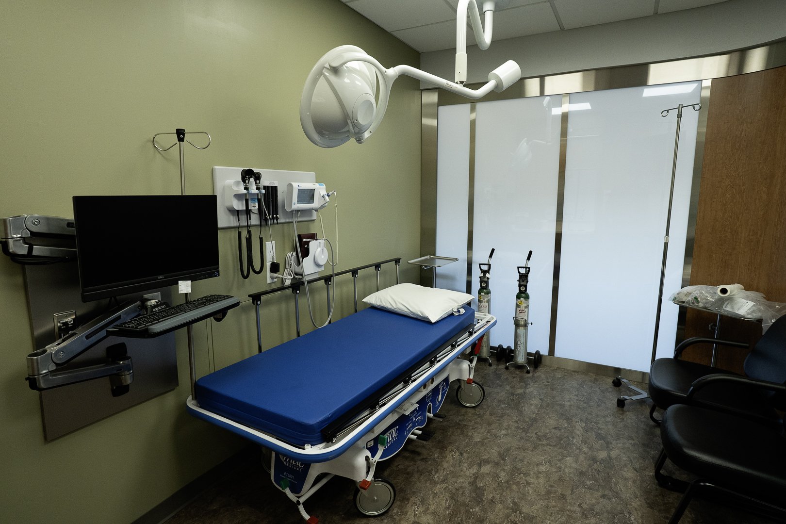 Hospital emergency room with a patient bed, medical monitors, oxygen tanks, an overhead surgical light, and chairs.