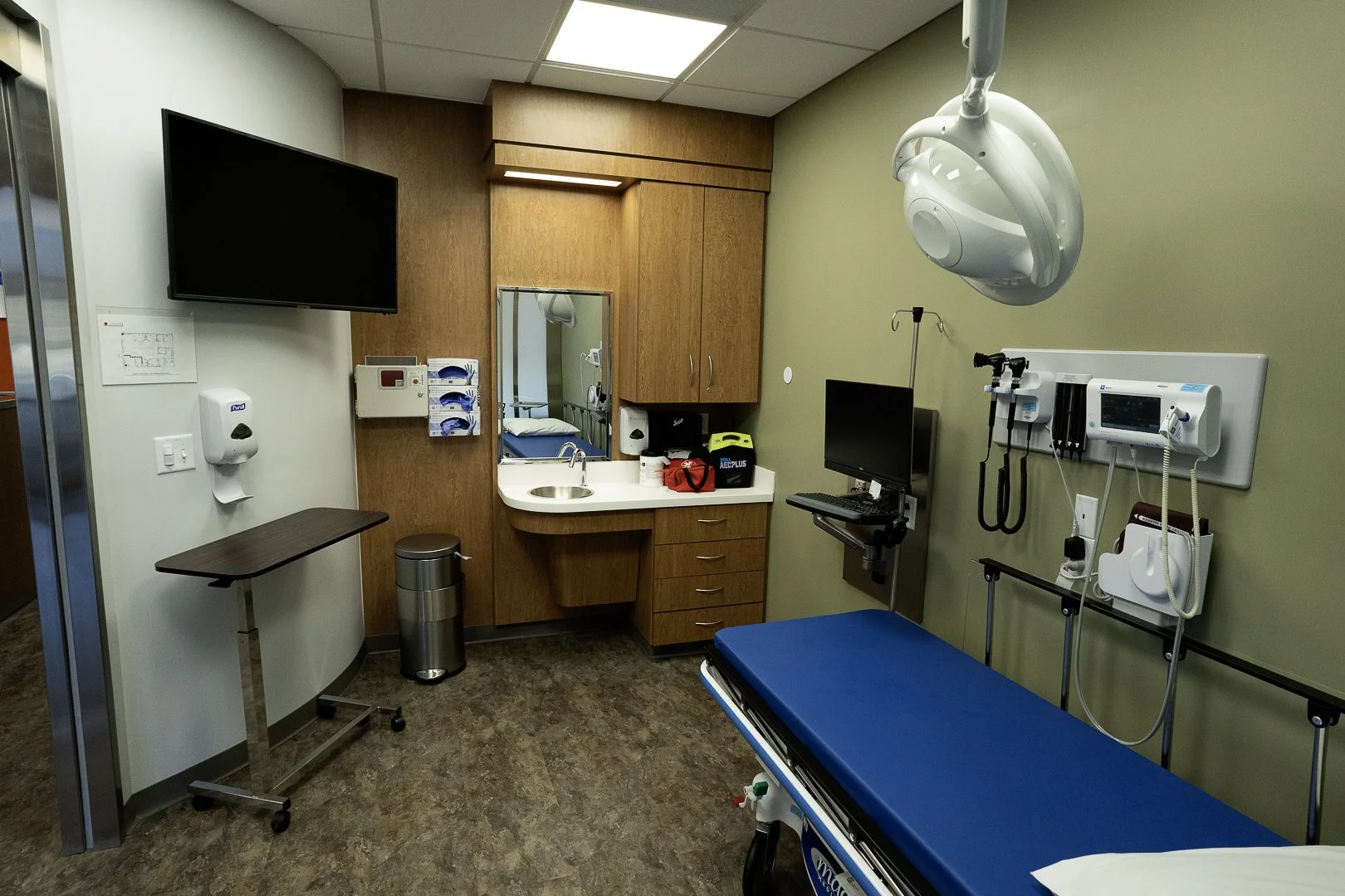 Empty hospital examination room with a hospital bed, medical equipment, monitors, and a small sink, with wooden cabinets and a wall-mounted TV.