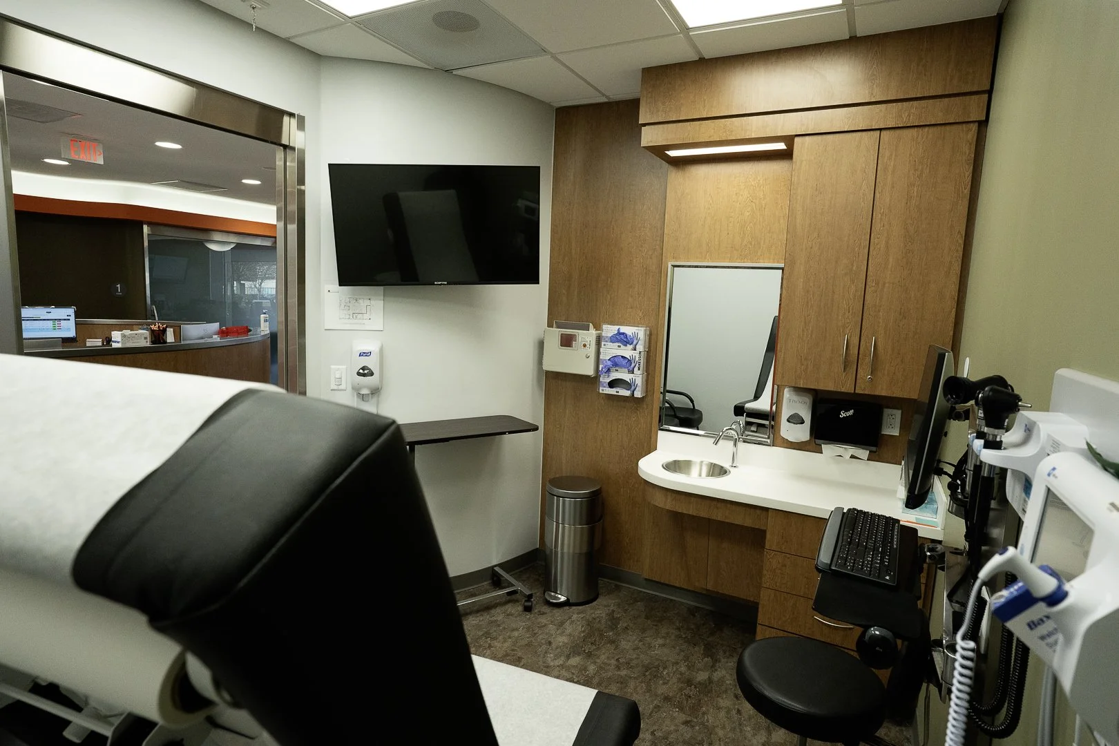 Medical examination room with a patient bed, wall-mounted TV, wooden cabinetry, computer equipment, and medical instruments.