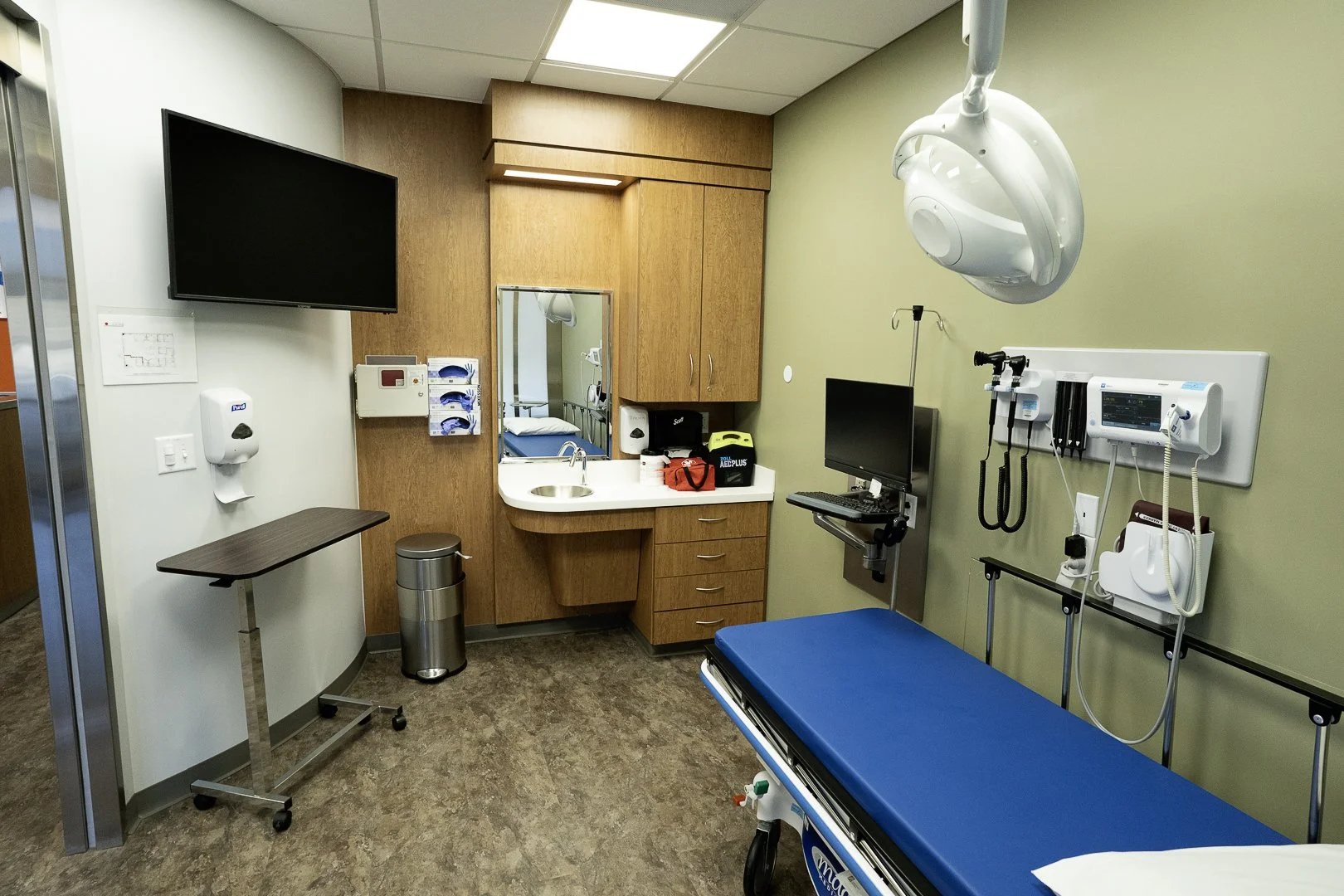 Empty hospital examination room with a blue examination table, a wall-mounted monitor, medical equipment, a sink area, and wood-paneled walls.