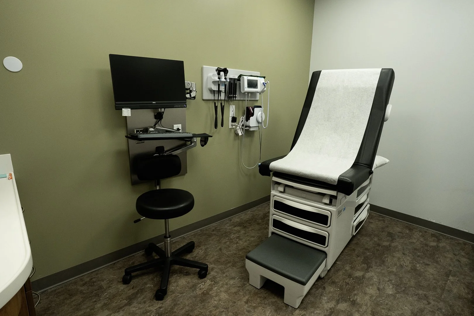 Medical examination room with a black-and-white exam table, a black stool, a wall-mounted monitor, and medical equipment on the wall.