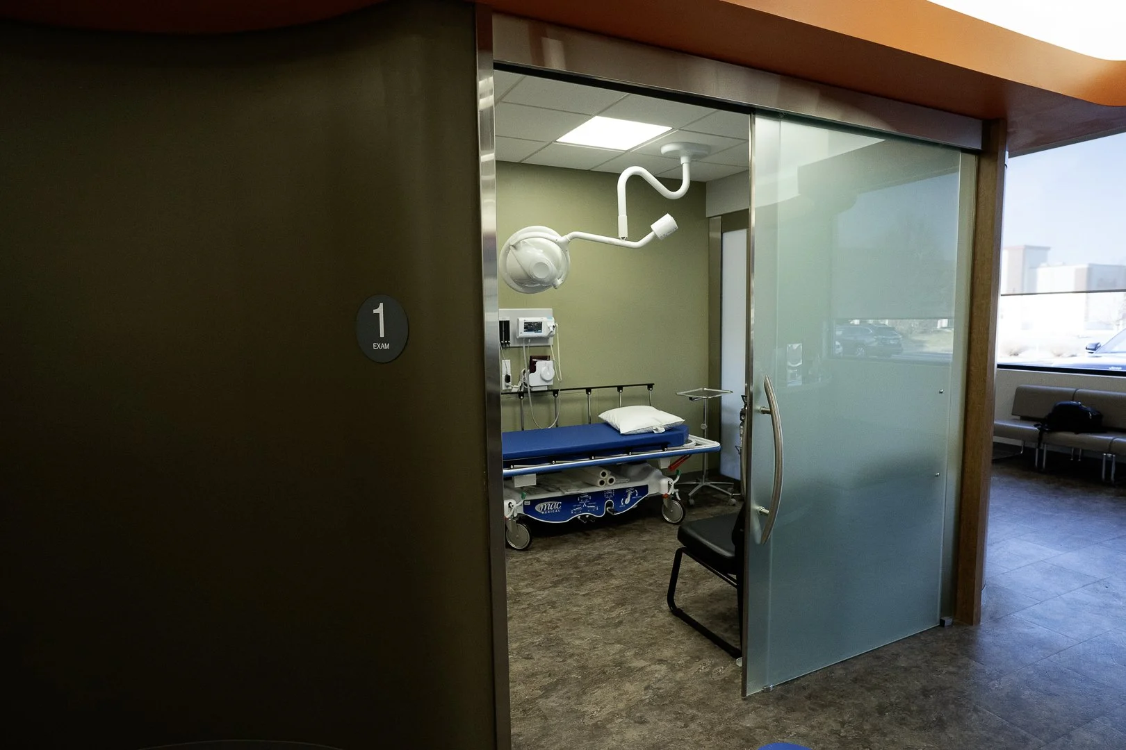 Empty hospital examination room, with an exam bed, medical equipment, and a chair, visible through a partially open glass door labeled '1 EXAM'.