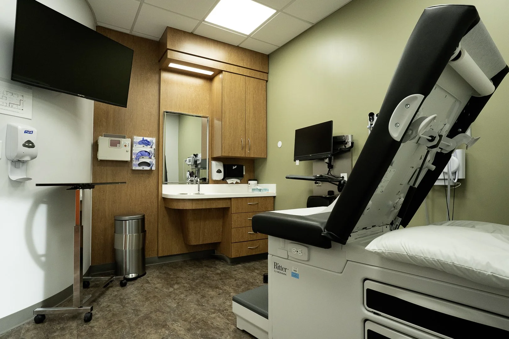 A medical examination room with a hospital bed, medical equipment, wall-mounted monitors, a sink with a mirror, and cabinets, with neutral-colored walls and flooring.