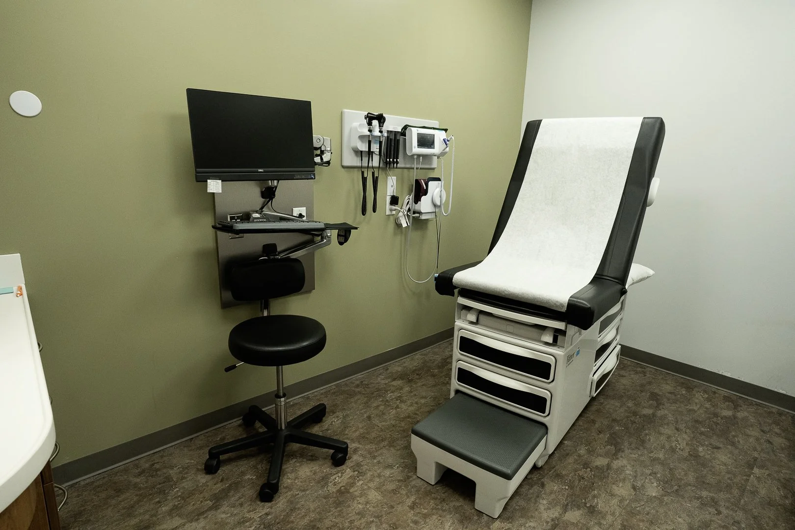Medical exam room with an examination table, wall-mounted medical equipment, and a computer workstation.