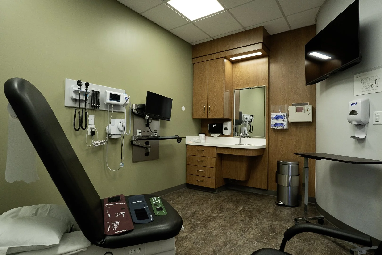 Medical examination room with an examination bed, medical equipment on the wall, a sink, cabinets, and a television.