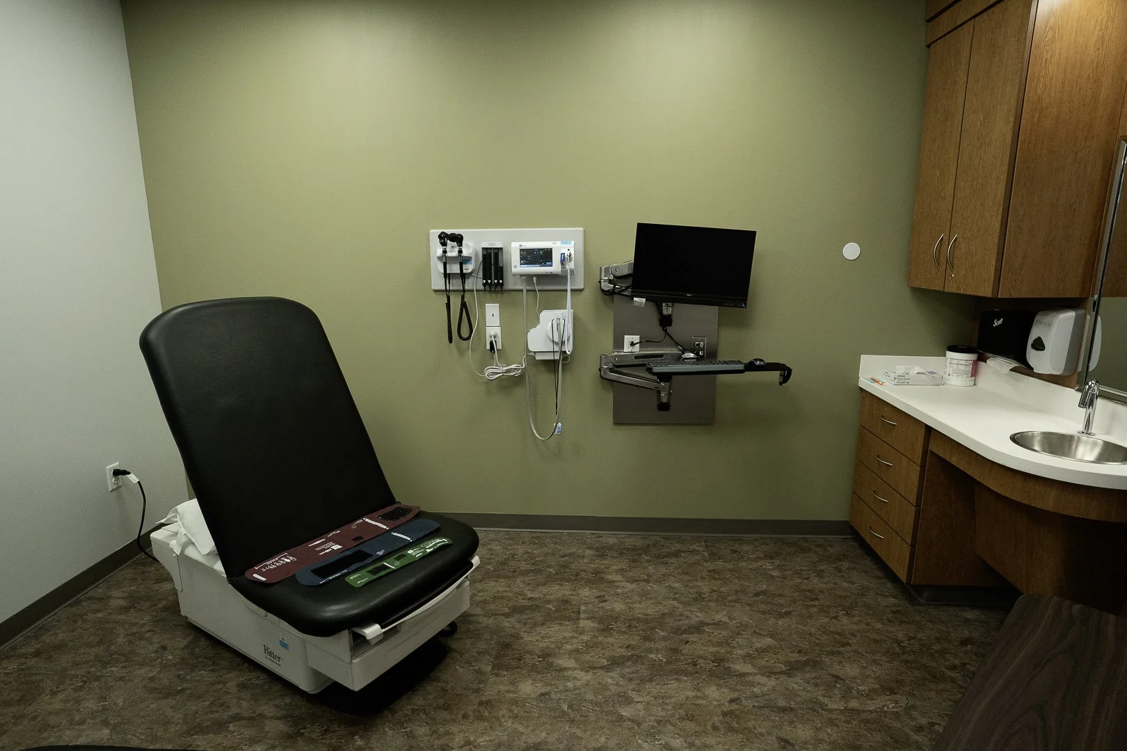 Medical examination room with exam chair, wall-mounted monitor and medical equipment, wooden cabinet, and sink.
