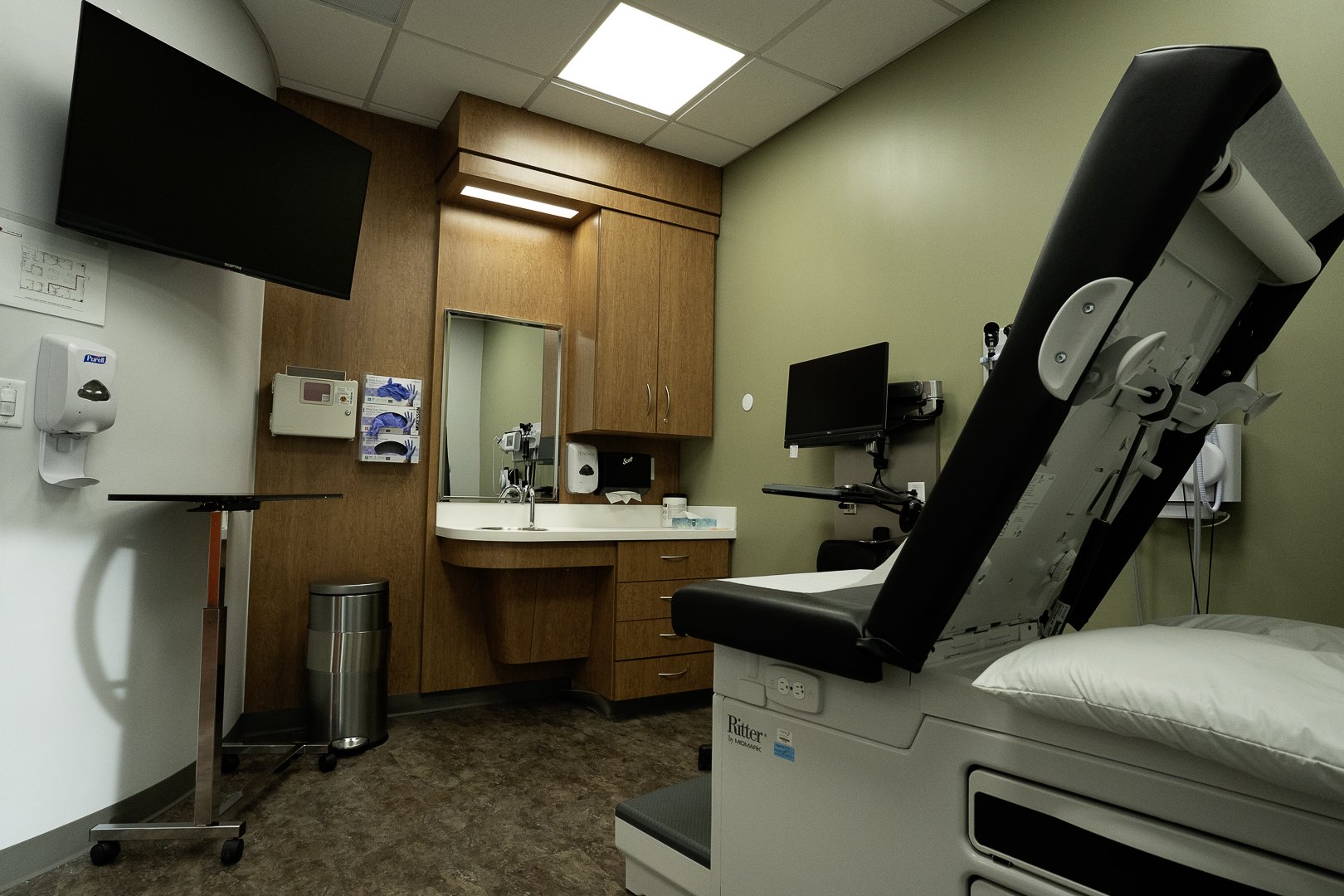 Hospital examination room with a hospital bed, medical monitors, a sink, a mirror, cabinets, and wall-mounted medical equipment.
