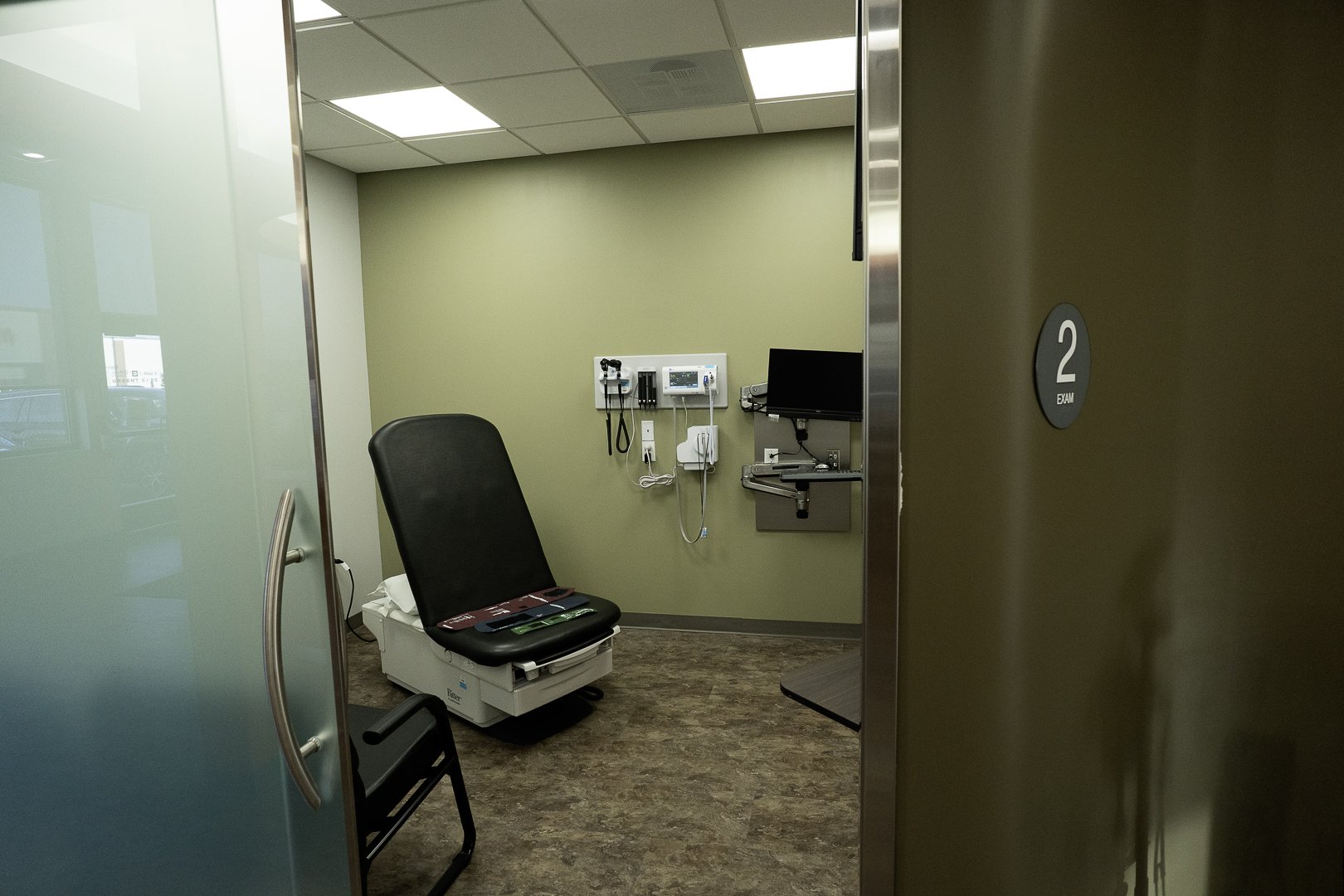 View of an examination room with a black medical chair, medical equipment on the wall, and a monitor, seen through a partially open door marked '2 Exam'.
