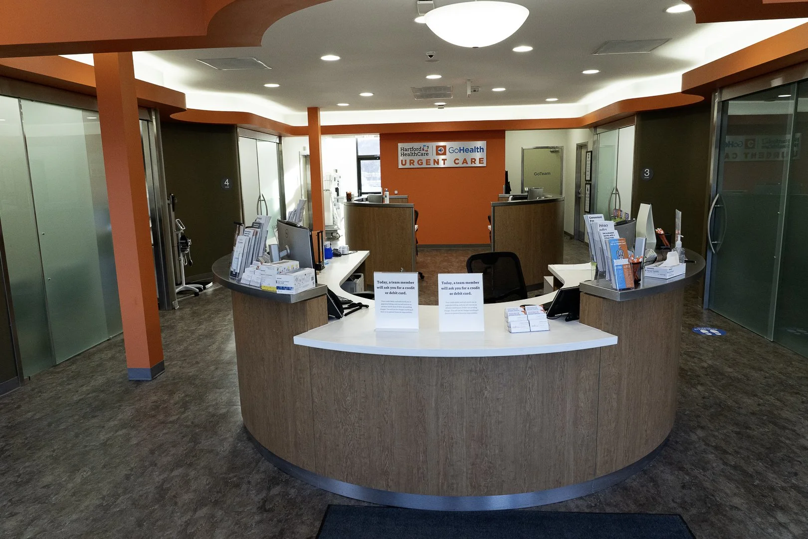Reception area of a Hartford HealthCare Urgent Care clinic with front desk counters, informational brochures, and signs on the desks, orange accent walls, and glass partitioned rooms.