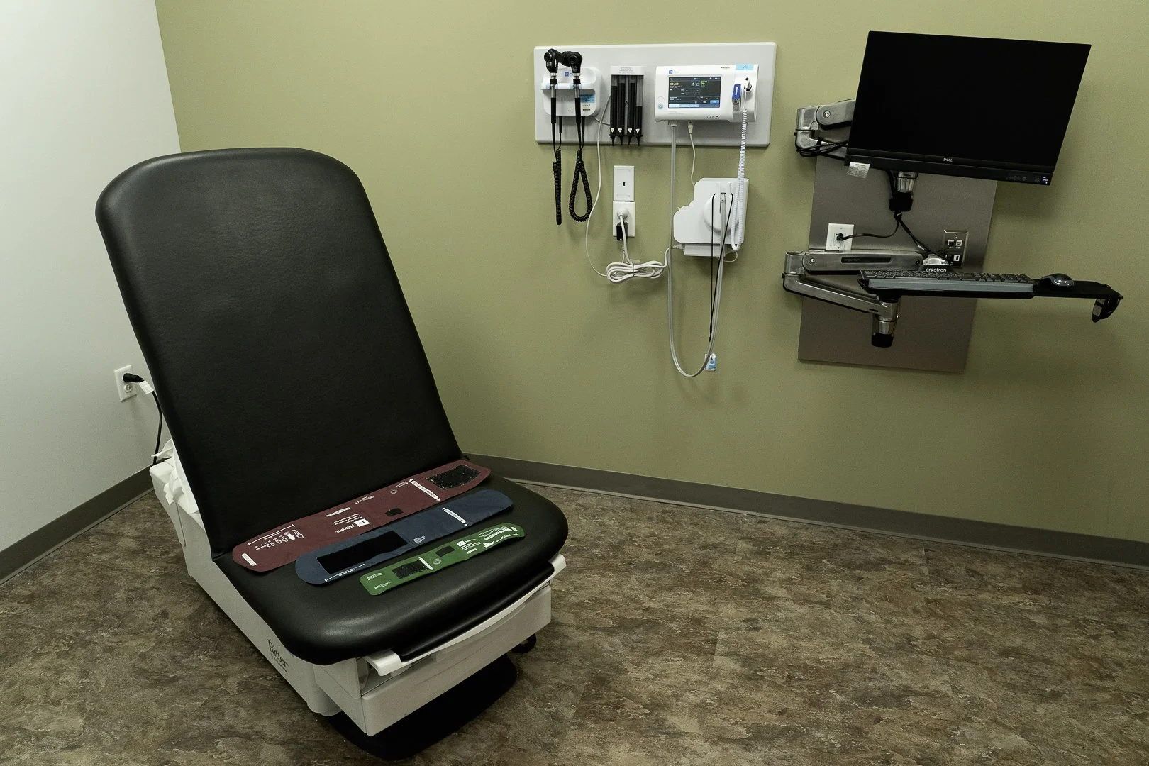 Exam room with medical examination chair, monitor, and medical equipment on wall.