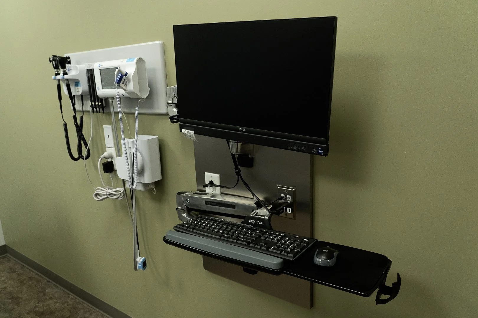 Hospital wall mounted medical equipment including a blood pressure monitor, otoscope, and thermometer, with a wall-mounted desk holding a computer keyboard and mouse.