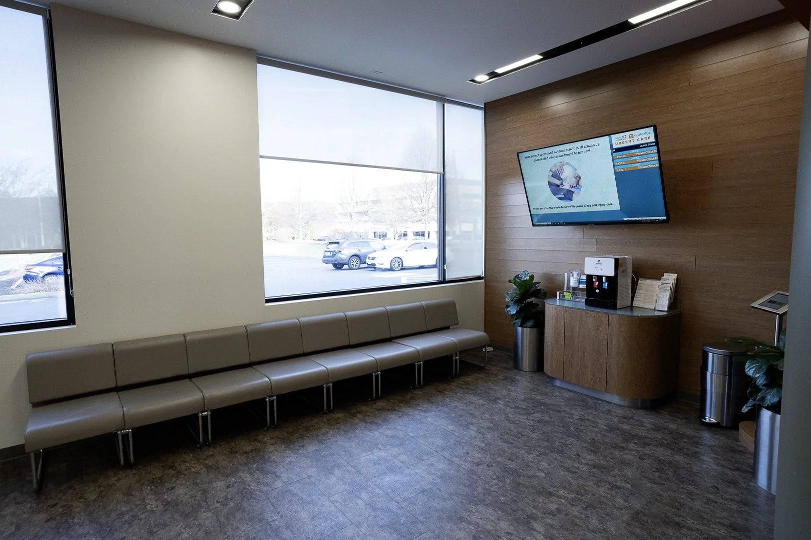 Medical waiting room with beige chairs against the wall, large windows with blinds, a wooden accent wall with a mounted TV screen, a small counter with a coffee machine and pamphlets, and potted plants.