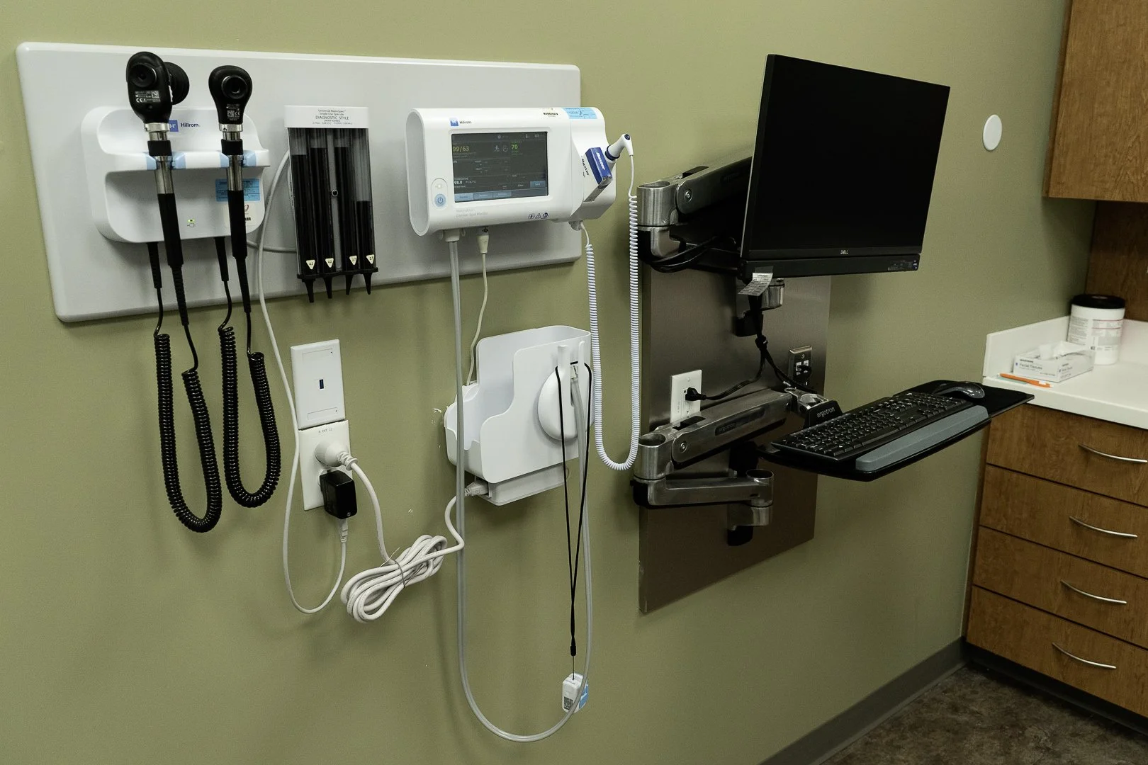 Medical examination room wall with diagnostic equipment including otoscope, ophthalmoscope, vital signs monitor, computer, and medical supplies.