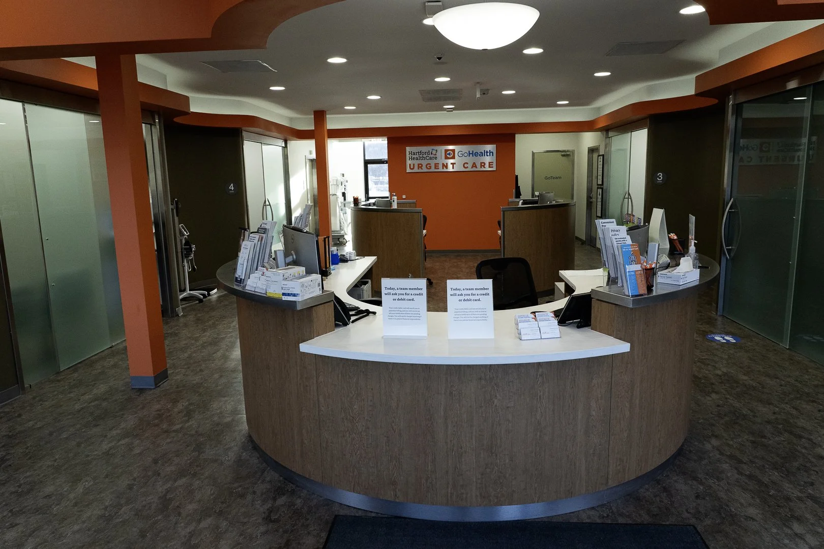 Interior of an urgent care clinic reception area with a curved wooden desk, computer monitors, informational pamphlets, and signs, with an orange accent wall and a sign indicating 'Hartford HealthCare GoHealth Urgent Care'.