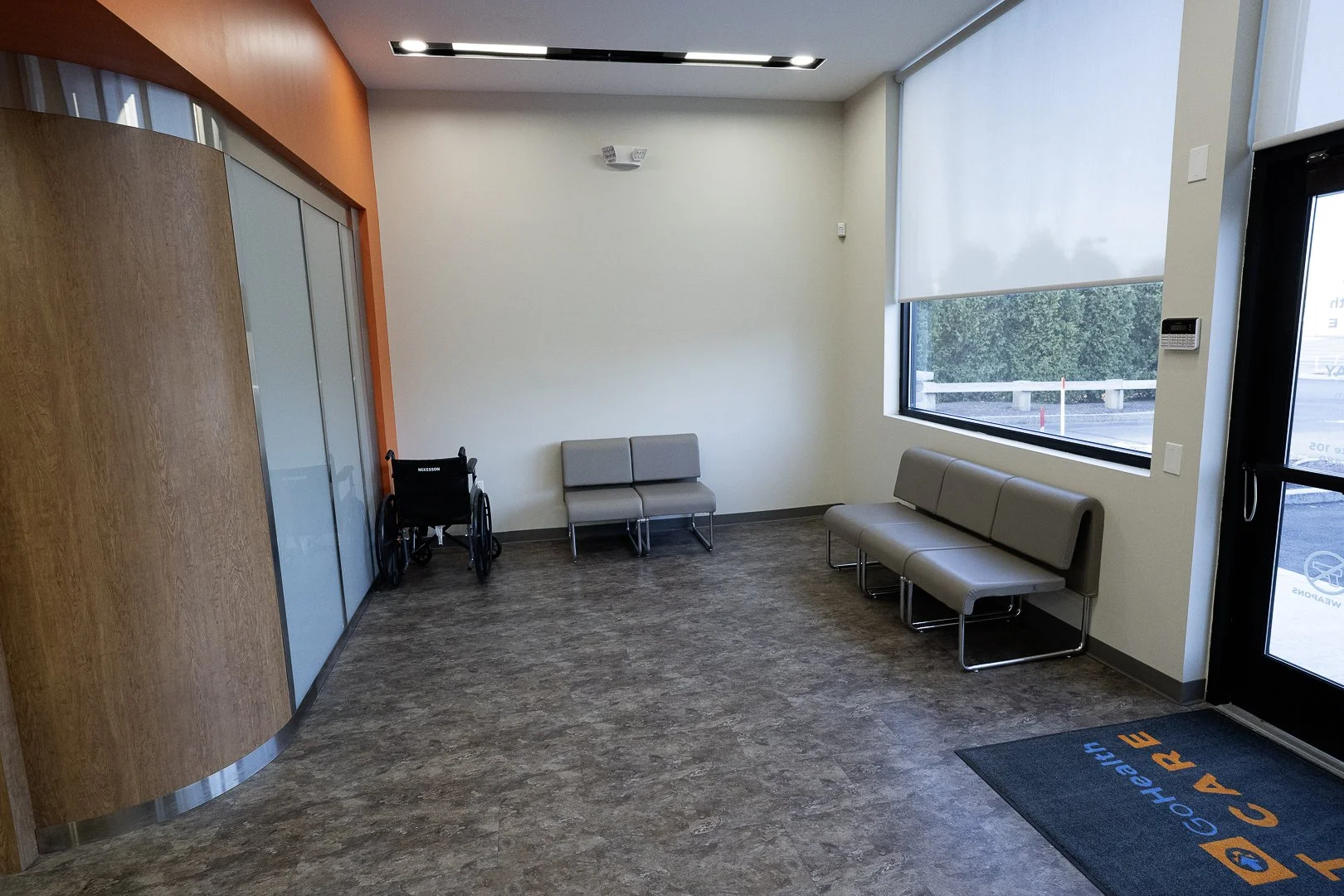 Waiting room with beige chairs, a wheelchair, large window with blinds, and a door with a welcome mat, in a medical office.