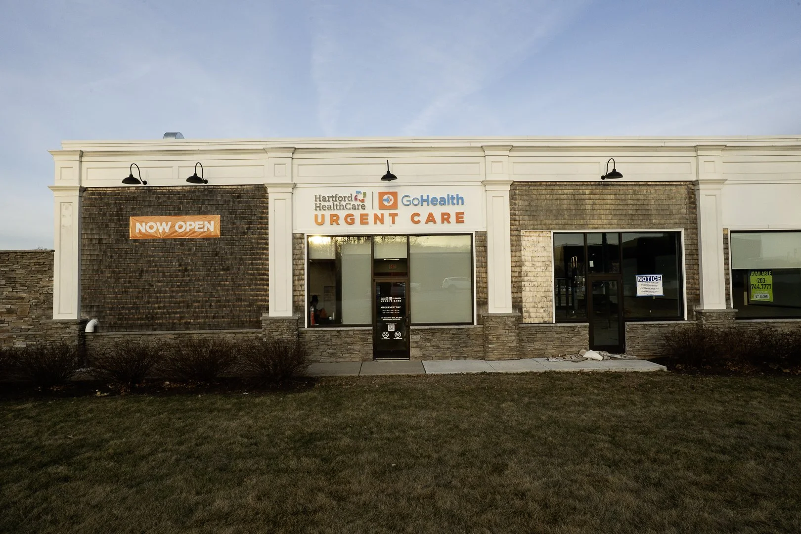 Front of a medical facility building with signs indicating it is now open, offering urgent care services, with a grassy area in front.