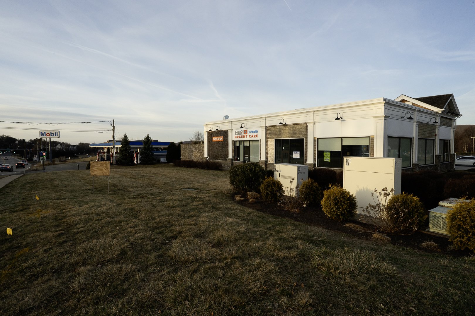 A commercial strip with a GoHealth Urgent Care center, a Mobil gas station, and a grassy area in front, during late afternoon with clear skies.