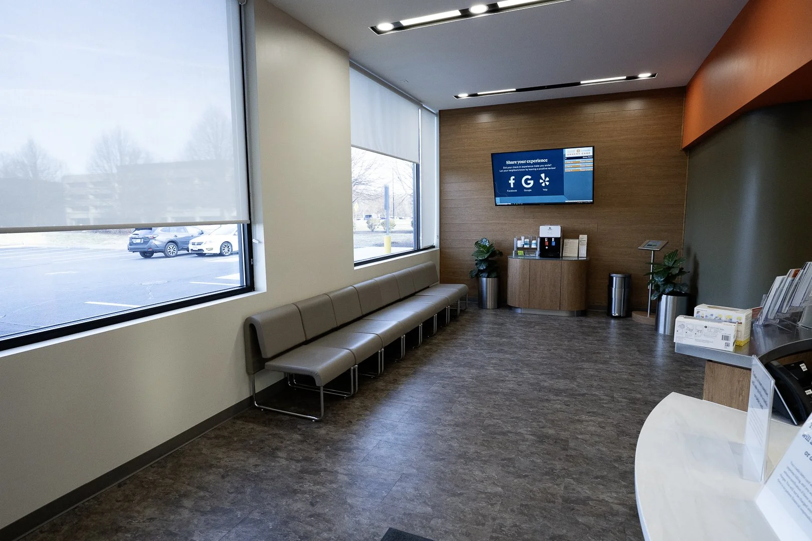Empty waiting room with beige chairs along the window, a wall-mounted television, and a small counter with a coffee machine and informational pamphlets.