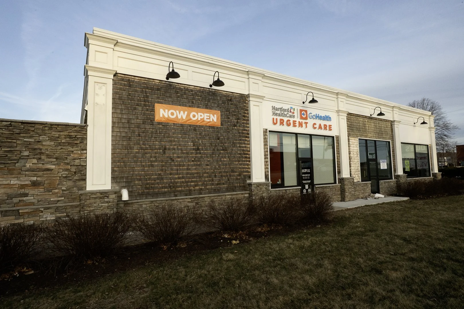 A single-story building with a sign that reads 'NOW OPEN' and advertising 'URGE NT CARE' for Hartford Healthcare and GoHealth. The building has large windows, a stone and siding exterior, and is surrounded by a lawn with some bushes.