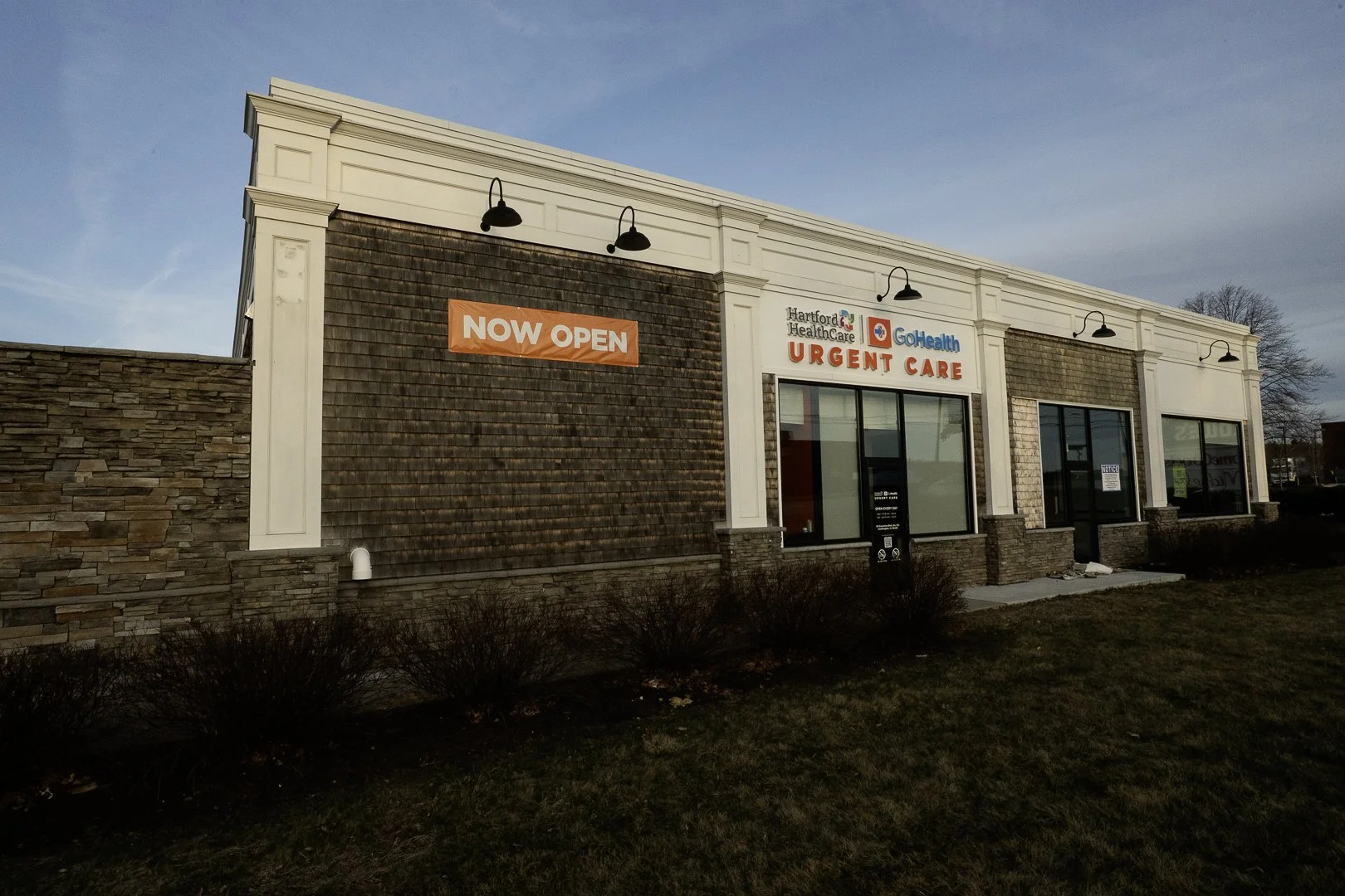 Exterior view of a medical building with signs for Hartford Healthcare GoHealth Urgent Care and a large orange banner that says 'NOW OPEN'.
