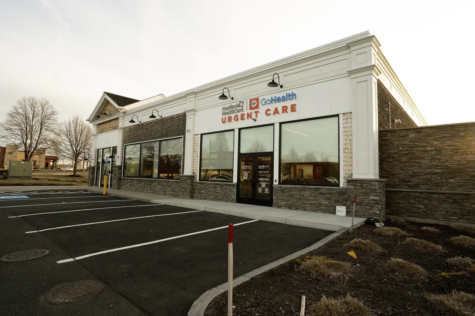 Exterior view of a medical clinic with a sign that reads 'Hartford HealthCare GoHealth Urgent Care,' a parking lot, and barren trees in the background.