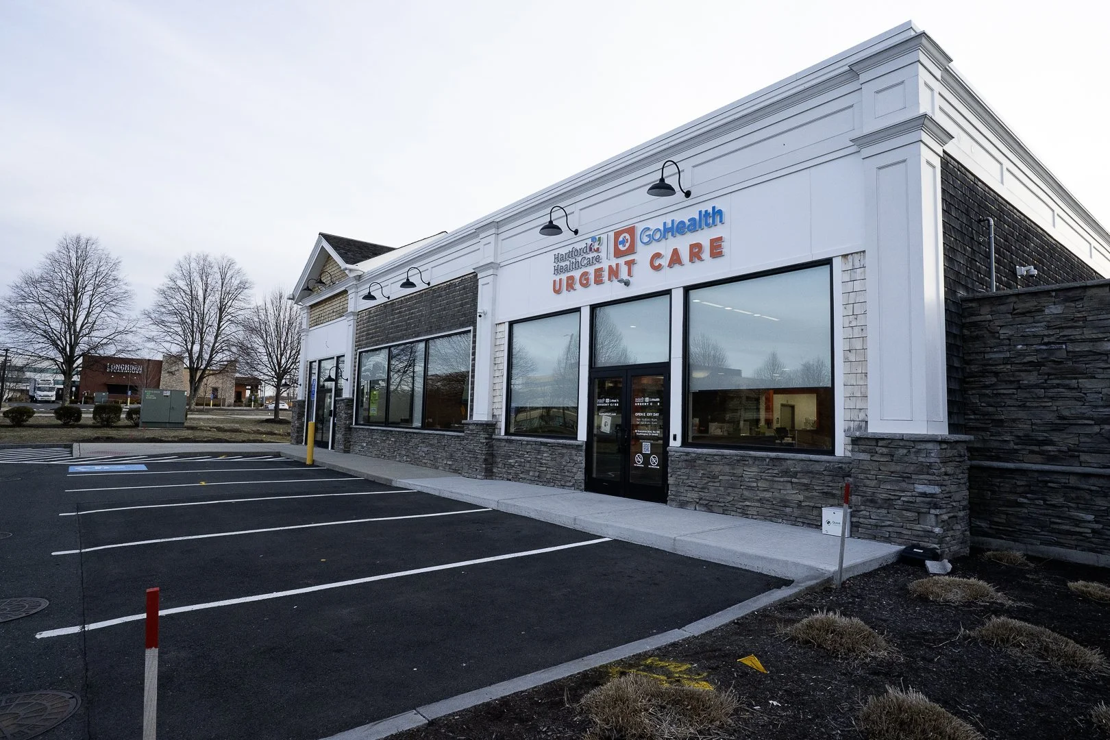 Exterior of a medical urgent care clinic with a sign reading 'Hartford HealthCare GoHealth Urgent Care', large glass windows, and an empty parking lot in front.